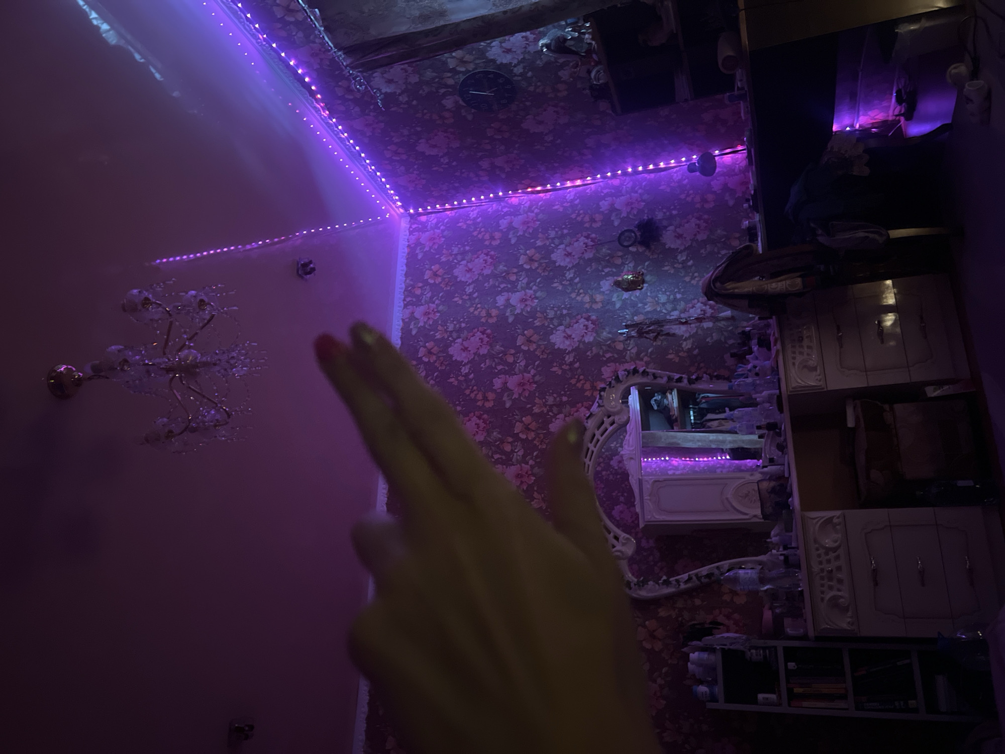 The image depicts a dimly lit bedroom with purple LED strip lights lining the ceiling and walls, creating a vibrant and atmospheric glow. A crystal chandelier hangs from the ceiling. In the foreground, a hand with painted fingernails is held up, partially obscuring the view. The background features a floral wallpaper, a vanity dresser with a large ornate mirror, and shelves filled with various items including books and bottles. A wall clock is visible on the right side of the room. The scene appears to be a personal space, possibly a teenager's bedroom, given the decorative lighting and the presence of personal belongings. The location, Krasnoe-na-Volge, Russia, suggests a specific context for the domestic setting.