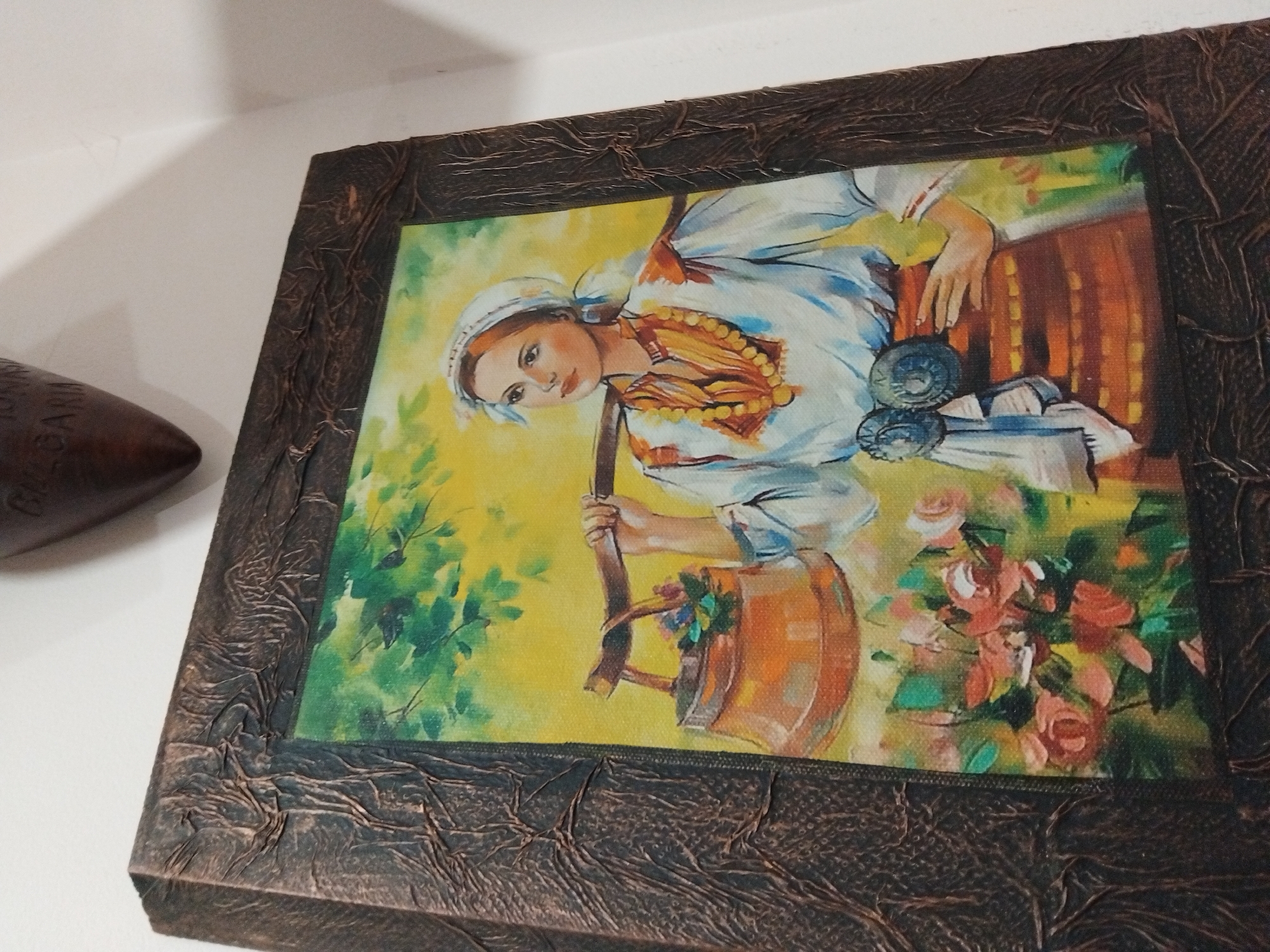 A close-up, angled shot shows a framed artwork hanging on a white wall. The artwork depicts a woman in traditional Bulgarian clothing, likely a folk art representation. She is shown carrying a bucket of water and is surrounded by greenery and flowers. The frame is dark brown with a textured, possibly leather-like, surface. To the left of the frame, a dark brown, rounded object with some illegible text ("BULGAR...") is partially visible. The location is possibly in Nova Zagora, Bulgaria, due to the artwork's style and suggested origin of the partially visible object.