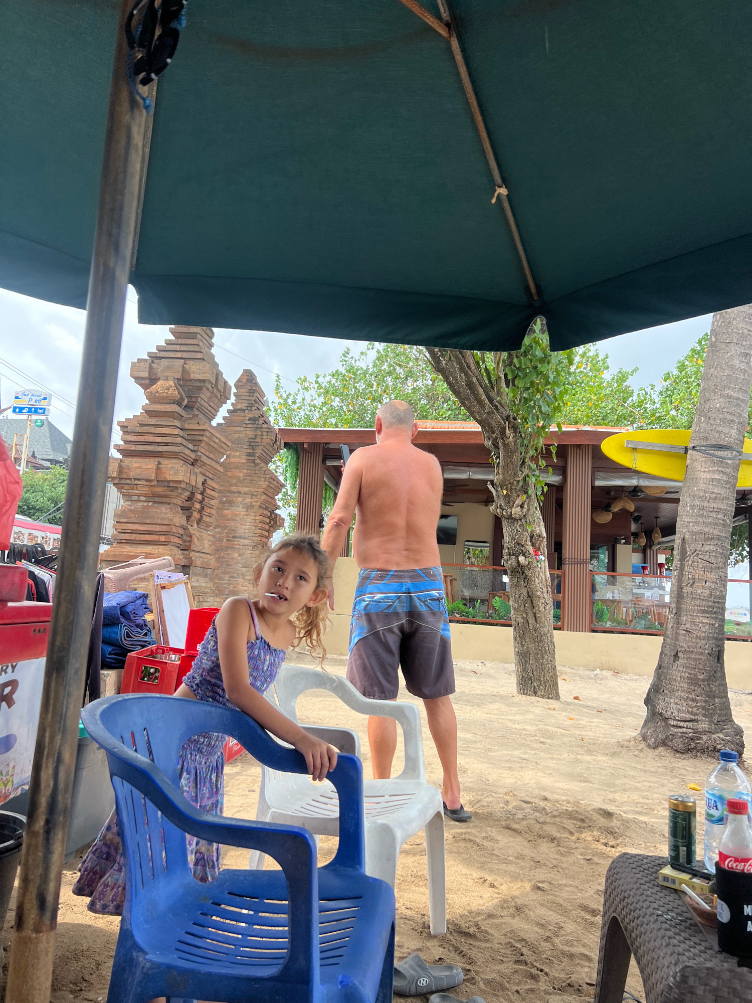 Underneath a large dark green umbrella on a sandy beach in Kuta, Indonesia, a young girl with light brown hair pulled back in a ponytail is seated on a blue plastic chair, looking directly at the camera with a lollipop in her mouth. She is wearing a purple and blue patterned sleeveless dress. Behind her, a shirtless man with short grey hair stands facing away from the camera, wearing blue and black patterned swim trunks. In the background, a tall, intricately carved brick structure, resembling Balinese temple architecture, stands prominently. To the right of the man, a building with a brown wooden exterior and a yellow surfboard displayed is visible. The ground is covered in sand, with scattered footprints. Several plastic chairs, a cooler, towels, and a red crate are present in the foreground. On a textured brown table to the right, a can of what appears to be a beverage, a bottle of Aqua water, a bottle of Coca-Cola, a pack of cigarettes, and a lighter are placed. The sky is overcast.