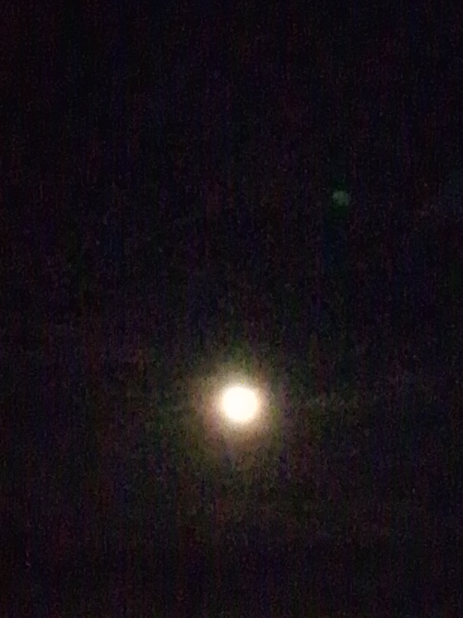 The image is a nighttime photograph of the sky. A bright, luminous object, likely the moon, is visible in the lower center of the frame, emitting a soft glow and surrounded by a faint halo. To the upper right of the moon, a smaller, greenish luminous object is present. The background is dark and appears to be a night sky with no discernible features like stars or clouds. The overall image is somewhat grainy, suggesting it may have been taken with limited light or at a distance. The scene is devoid of any human presence or man-made structures. There is no information in the image to indicate its location in Serti, Nigeria.