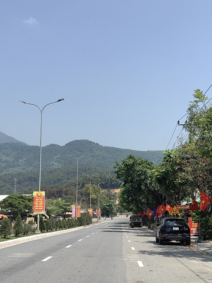 The scene depicts a wide, paved road in Hải Vân Ward, Vietnam, extending into the distance with forested mountains in the background under a clear blue sky. The asphalt road features two visible lanes separated by white dashed lines, with a median strip to the left containing rows of small evergreen trees and multiple tall streetlights equipped with dual LED fixtures. Large red banners with yellow Vietnamese text and the yellow star emblem are displayed on poles along the median. The right side of the road is bordered by dense green foliage, from which numerous small red Vietnamese flags with a yellow star are strung, accompanied by yellow triangular warning signs. A black SUV is parked on the dirt shoulder on the right, facing away from the camera, and further down the same side, a dark-colored utility truck is also parked. Two motorcycles are visible in the far distance on the road. Overhead utility lines traverse the upper right of the frame. No individuals are distinctly visible or engaged in specific actions or interactions.