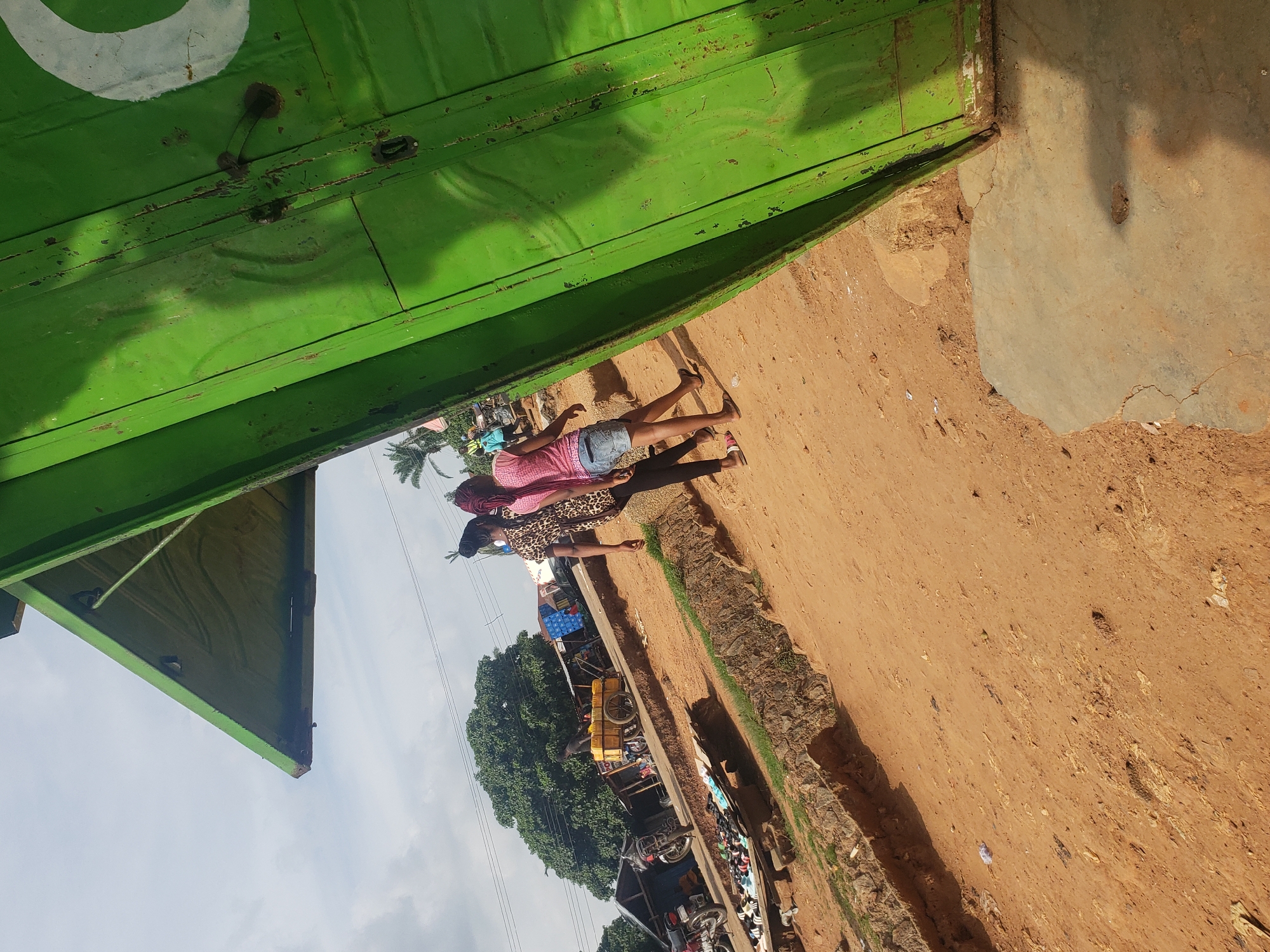 The image depicts an outdoor scene in Kachia, Nigeria, likely a marketplace area. In the foreground, a large, weathered green structure, possibly a stall or shop, dominates the right side of the frame, with a white curved marking visible on its surface. To the left of this structure, two young women are walking away from the viewer. The woman on the left wears a leopard print top and black pants, while the woman on the right is in a pink t-shirt and denim shorts. The ground is predominantly dry, reddish-brown dirt, with some scattered debris. Behind the women, a row of market stalls can be seen, some with roofs. Various objects are visible at the stalls, including what appears to be a yellow cart with goods, motorcycles, and a collection of footwear displayed on the ground. The sky above is partly cloudy. The overall impression is of a typical busy market environment in a developing urban setting.