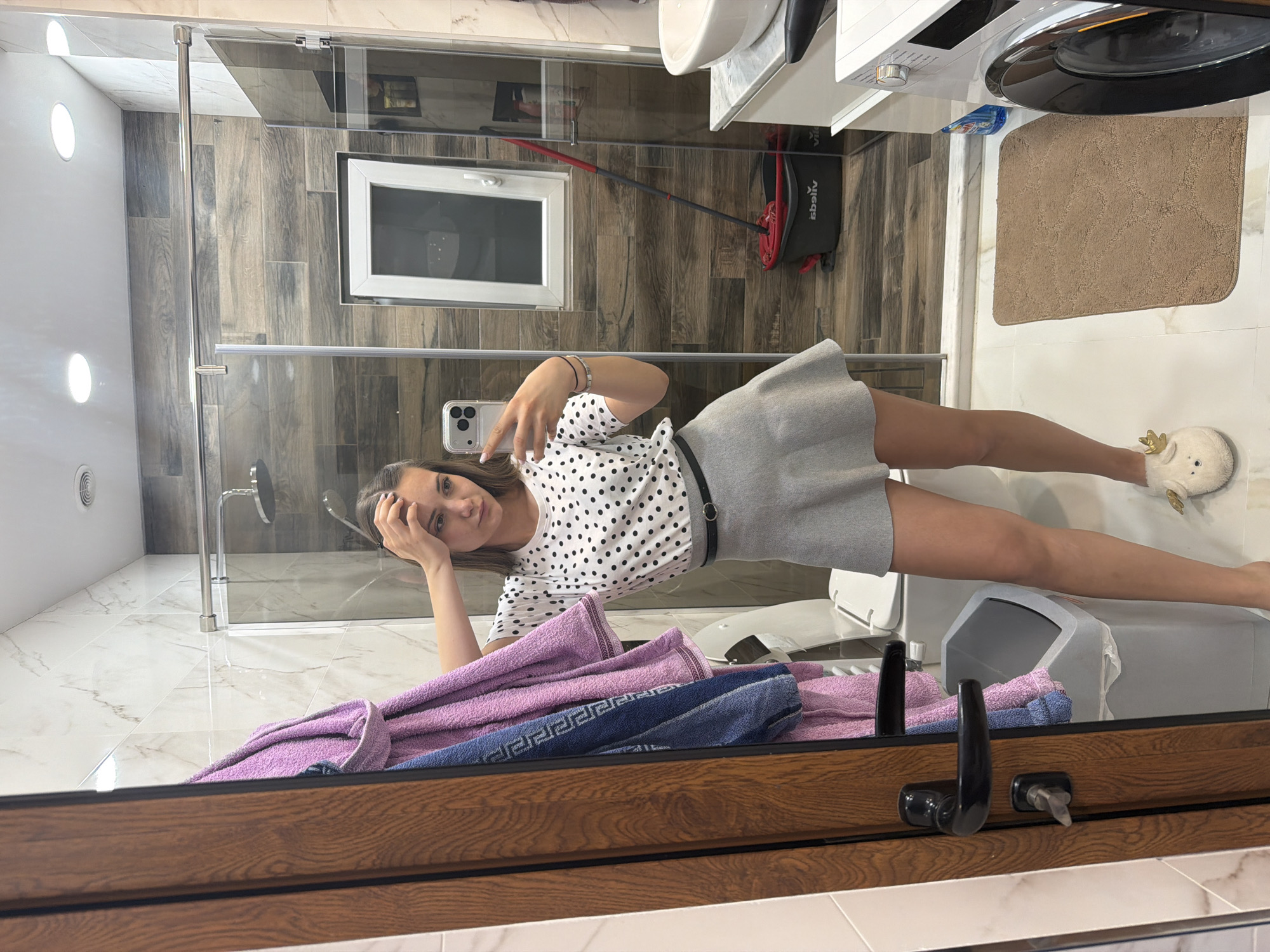 A young woman is seen in a bathroom reflected in a mirror, holding her phone and taking a selfie. She's wearing a white polka dot top, a grey skirt with a black belt, and white slippers with a decorative bunny face. The bathroom features a shower, a washing machine, a sink, and a bath mat. There are purple and blue towels near the bottom of the frame, close to the mirror. The bathroom appears to be located in a private residence. There is no way to independently verify that this image was taken in Buhovo, Bulgaria.