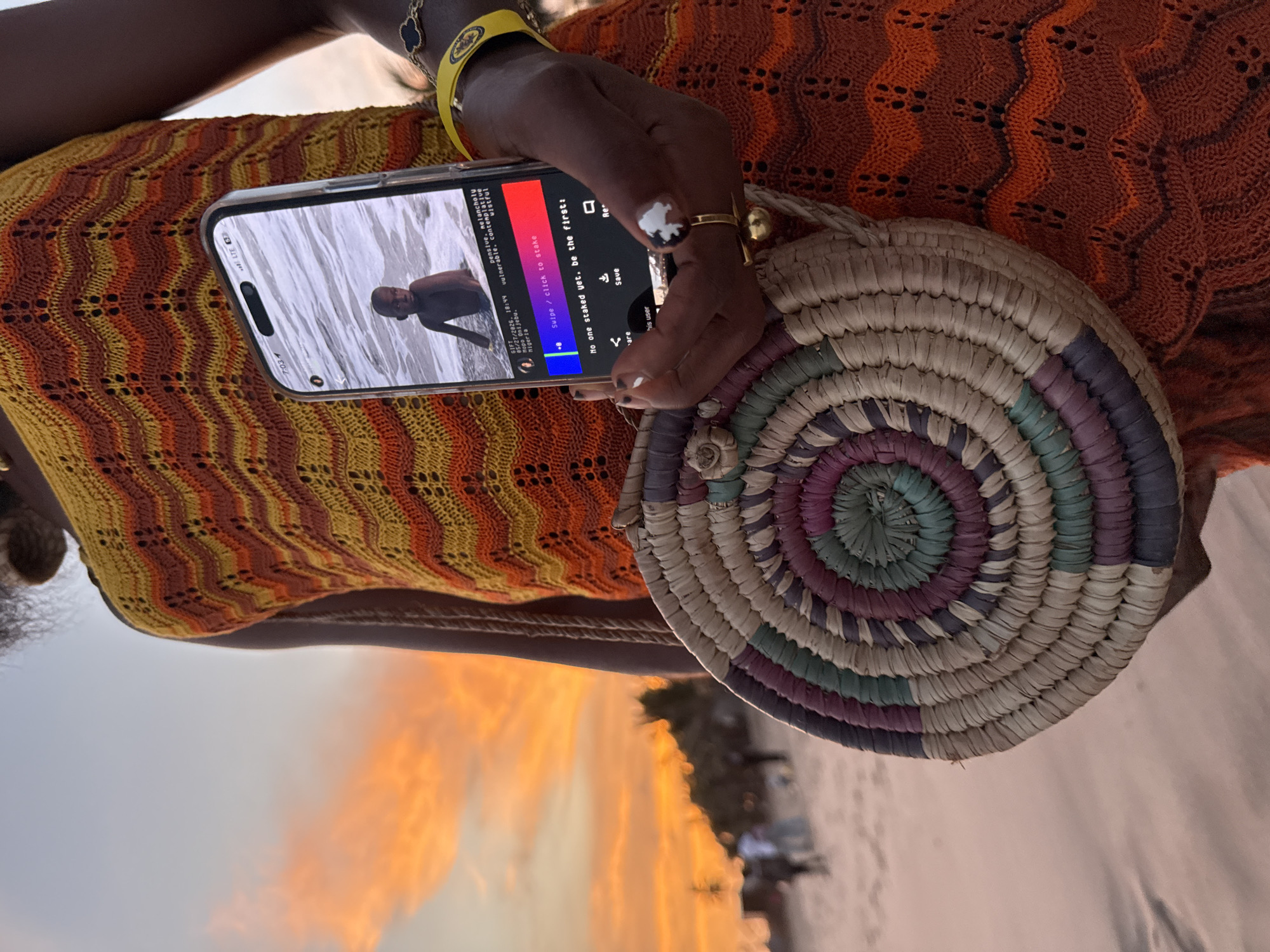 A person is holding a phone displaying a photo of a shirtless person in water. The phone screen also shows text related to staking and other information. The person holding the phone is wearing rings and a yellow bracelet. They are also holding a circular woven bag with multicolored stripes. They are wearing an orange and yellow dress. The background shows a beach scene with an orange and yellow sunset. The image may be taken on a beach in or near the city of Mopo Onijebu, Nigeria, based on the text on the phone screen.