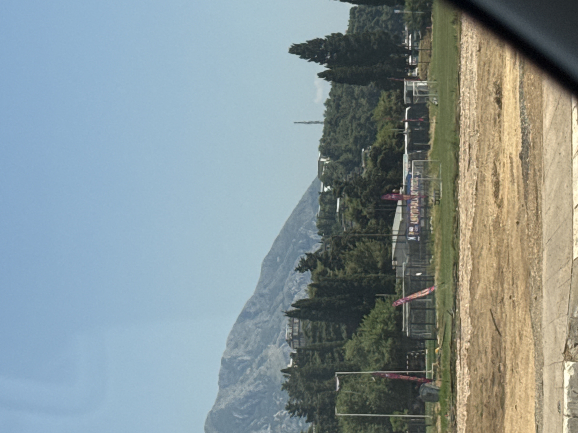 This is a photograph of a sports field and a mountainous landscape, likely taken in Budva, Montenegro. The image is oriented vertically, with the sky at the top and the ground at the bottom.
The foreground shows a dirt area and a paved path, suggesting a construction site or an unpaved section of the sports facility. Beyond this, a green grass field is visible, with several white-framed goals scattered across it, indicating a football or soccer pitch. There are also some scattered trees and buildings in the middle ground. In the background, a large, rugged mountain dominates the scene. The sky is a clear, pale blue, suggesting a sunny day. The overall impression is one of an outdoor recreational area nestled against a natural, mountainous backdrop.
The time of day appears to be daytime, possibly late morning or early afternoon, given the bright sunlight and clear sky. There is no discernible activity or event taking place in the image. The setting appears to be a sports complex or training ground, with the imposing mountain range providing a dramatic natural backdrop. Some of the structures in the middle ground have visible text, including what appears to be "PLAZA" on one of them. The weather is clear and sunny.