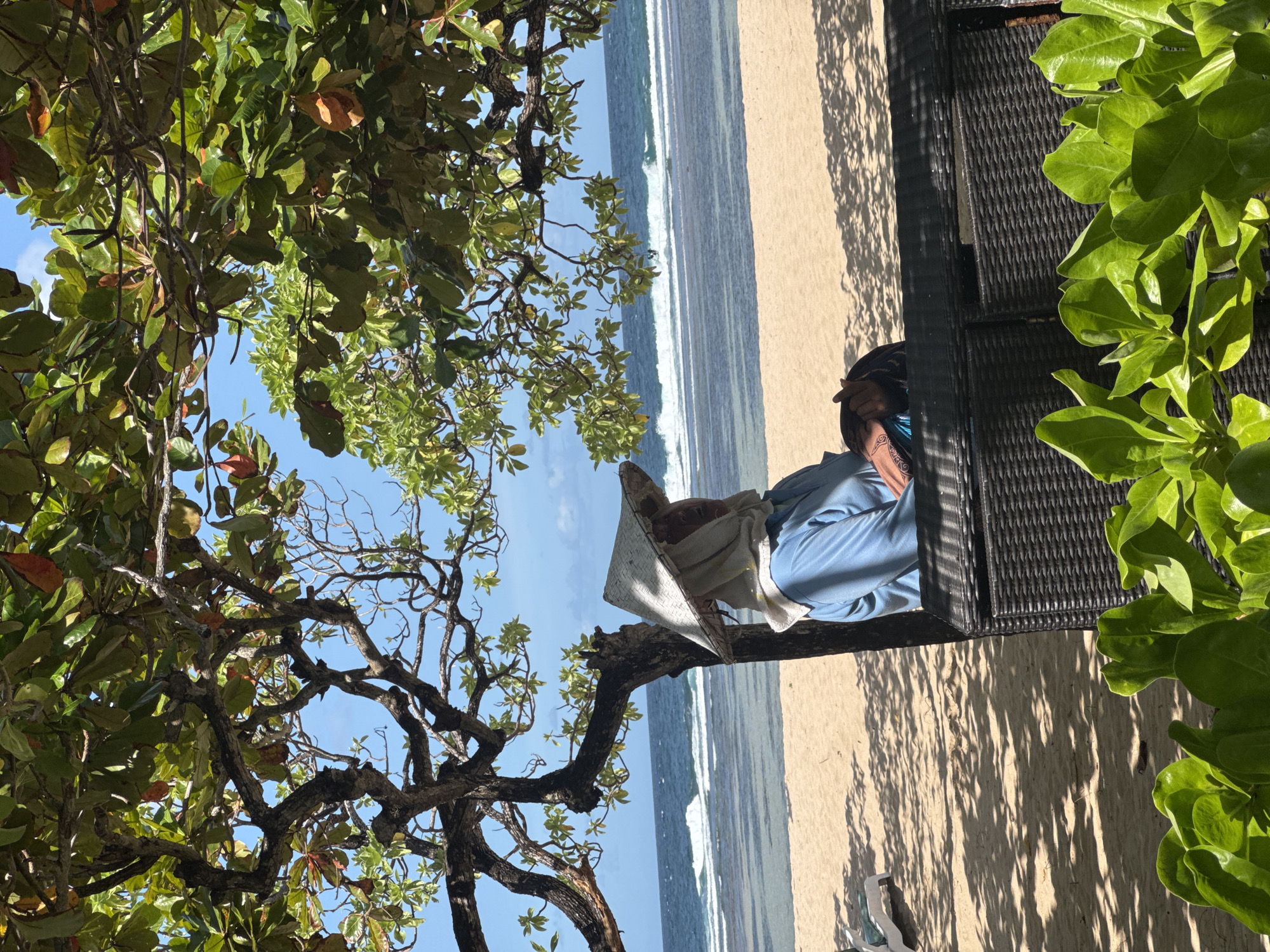 The image shows a person sitting on a chair on a beach, likely in Kuta Selatan, Indonesia, given the coastal environment. The person is wearing a conical hat and a blue shirt. The beach has light brown sand and blue water with white waves. Foliage from trees frames the view, appearing on the left and right sides of the image. The photograph is taken from a slightly elevated perspective, looking down towards the subject and the beach.
