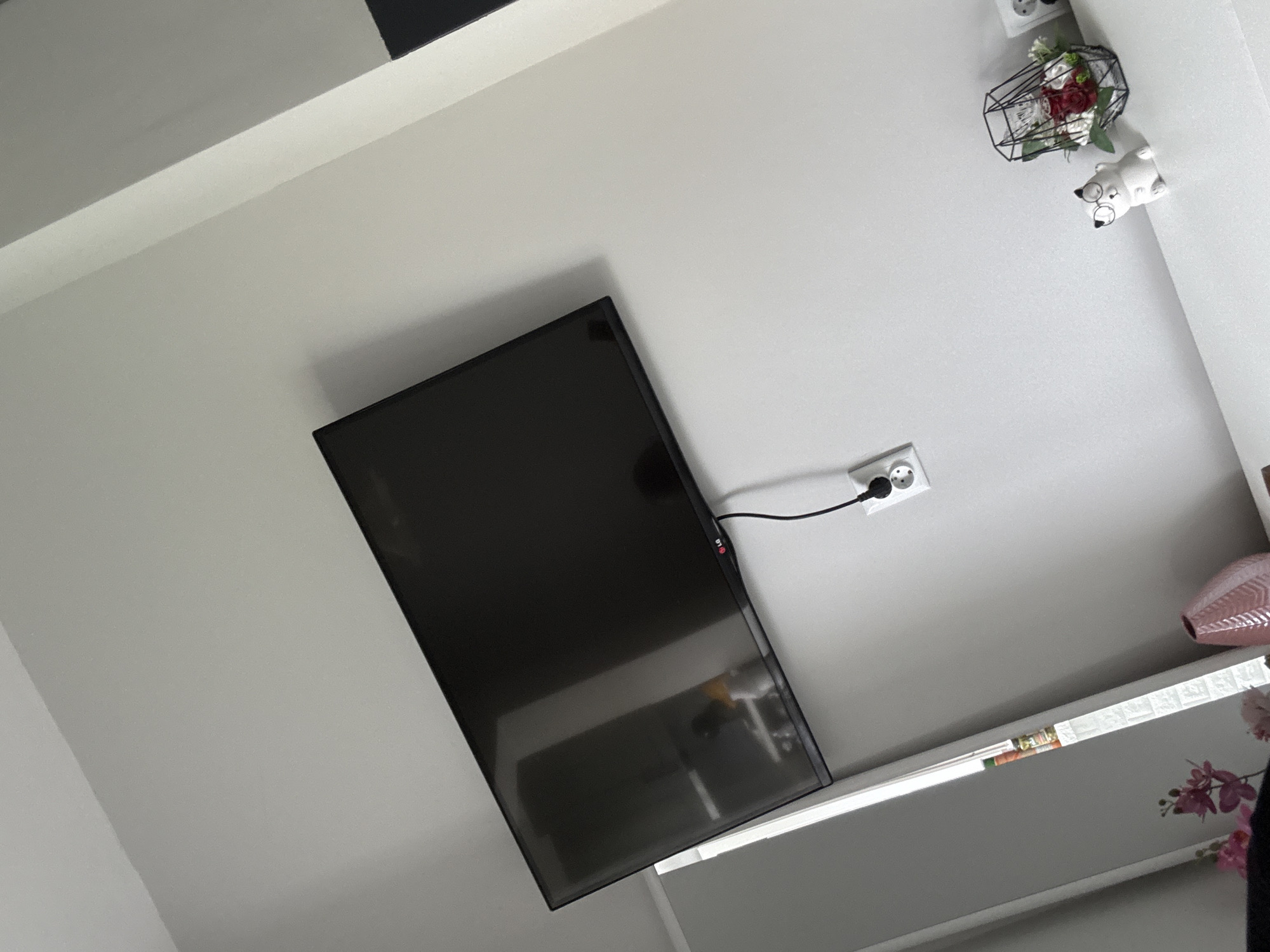 The image displays an interior room from a tilted, upward-looking perspective, focusing on a wall-mounted entertainment setup and decorative elements. A large, dark flat-screen television, with the "LG" brand visible on its bottom right bezel, is prominently featured. It is connected to a white electrical outlet via a black power cord.
Below the television, a long, light-colored shelf or media console is visible. On this surface, a book or magazine with the word "Bank" clearly legible on its spine can be seen, alongside some artificial pink orchid flowers on the far right. Partially visible on the right edge is a textured pink vase.
Above and to the right of the television, several decorative items are arranged against the light grey wall. These include a black wire cage-like ornament filled with artificial red and white roses and green foliage. Next to it, a small, white ceramic figurine, resembling a pig or dog with distinct snout and eye features (possibly glasses), adds a whimsical touch. Another electrical outlet is partially visible further up on the wall.
The walls and ceiling are painted in a uniform light grey or white tone, creating a bright and clean aesthetic. The lighting suggests a well-illuminated indoor environment, likely daytime or under bright artificial lights. No people or specific activities are present, the scene is static, showcasing room decor. The location context for this interior scene is Stara Zagora, Bulgaria.