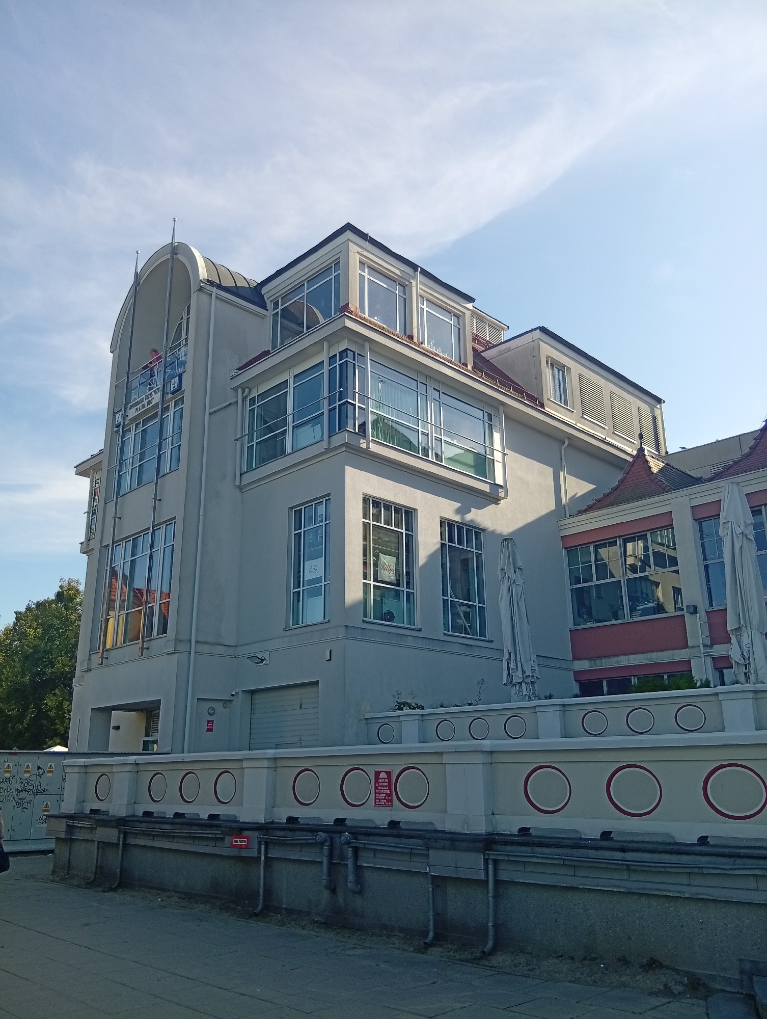 The image shows a building in Sopot, Poland, with a modern architectural style featuring large windows and a light-colored facade. The building appears to be multi-story with several levels, including attic windows. In front of the building is a low wall decorated with circular patterns. The sky is blue with scattered white clouds. Trees are visible to the left of the building. There are also some closed umbrellas in front of the building. Some signs are visible on the wall and on the building itself. The overall scene suggests a sunny day.