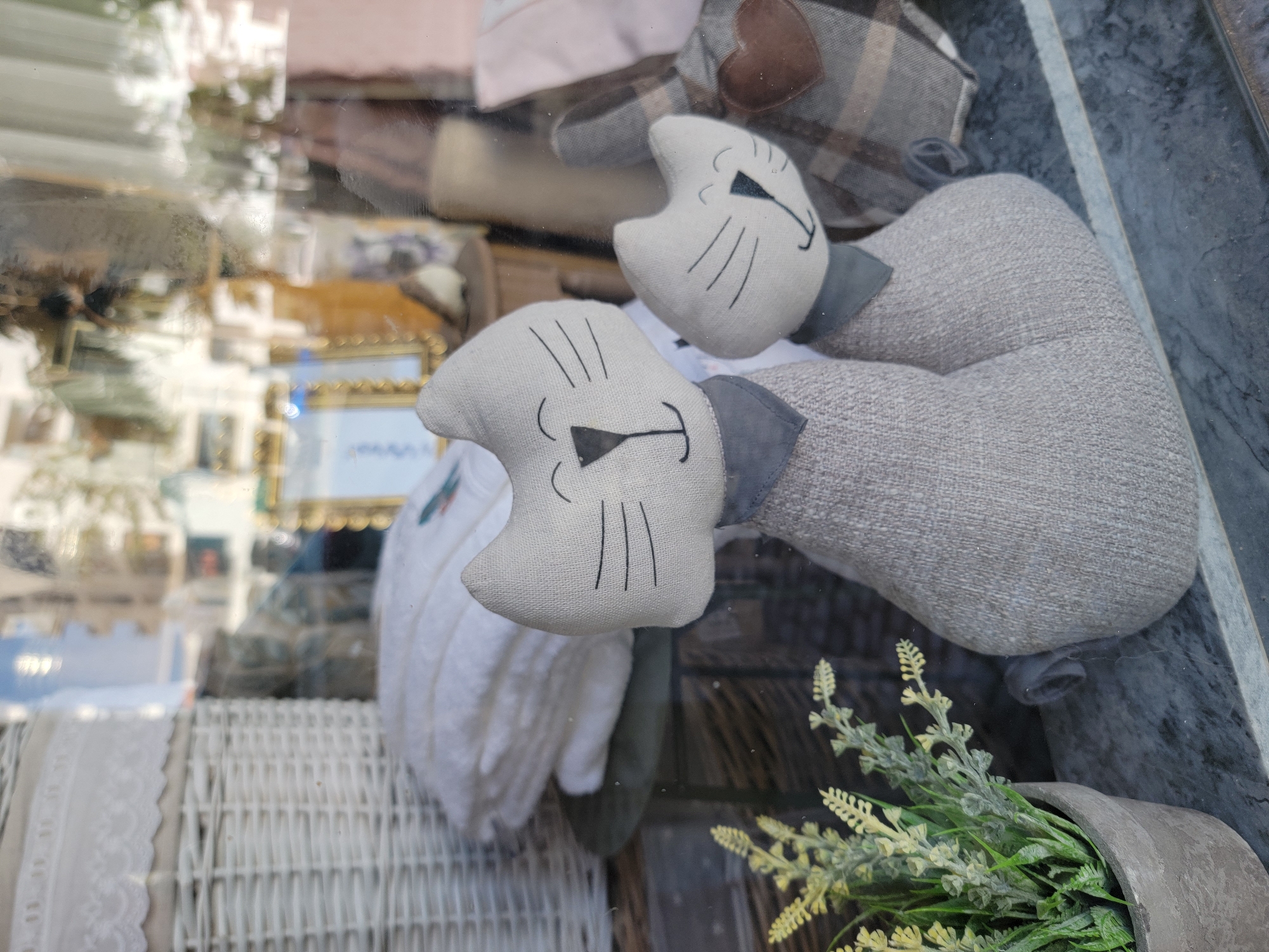 In Quarteira, Portugal, a storefront window displays a collection of decorative items. The foreground features two fabric cat figurines, a larger one in front and a smaller one behind it. Both cats are a light grey color and have embroidered facial features, including black lines for whiskers and mouths, and simple triangular noses. They also wear grey fabric bandanas. To the left, a potted plant with green foliage and small yellow flowers adds a touch of nature to the scene. Behind the cats, stacks of white folded fabric, possibly towels or linens, are visible, along with a white woven basket. The reflection in the window shows a blurred view of buildings and trees. The overall presentation suggests a retail environment, likely a home decor or gift shop.