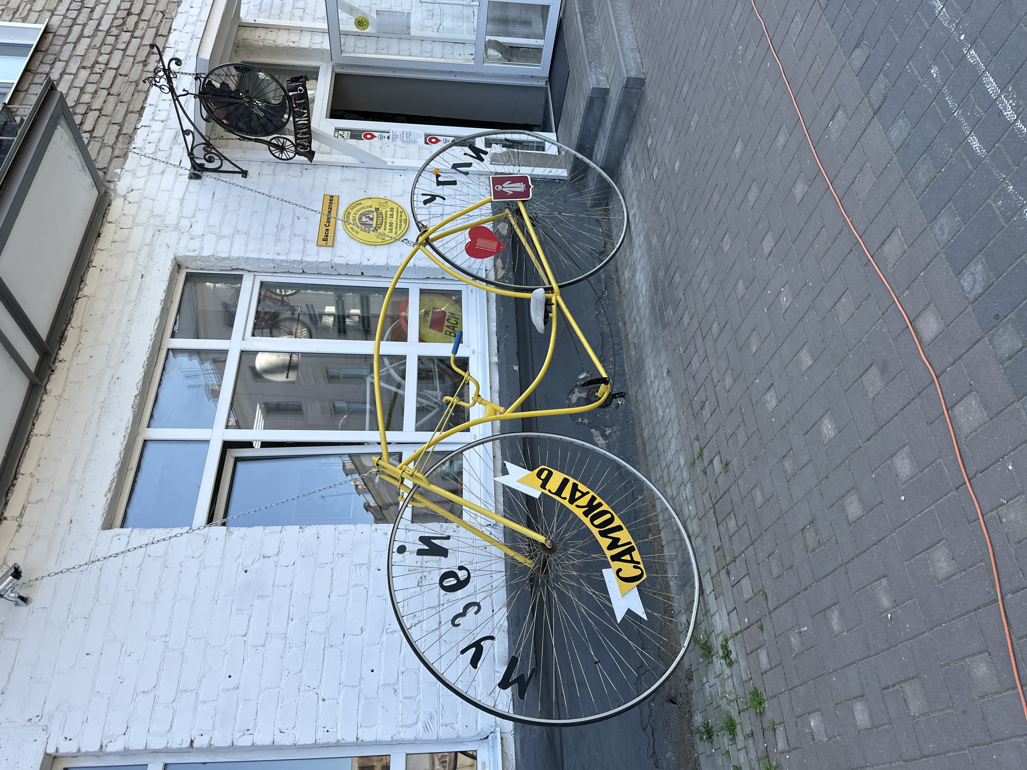 A large, yellow, novelty bicycle sculpture is displayed outside a building in Nikul'skoe (Neverkovskii S/O), Russia. The bicycle has extremely oversized wheels, with the front wheel significantly larger than the rear. Black lettering is painted on the spokes of the front wheel, reading "МУЗЕЙ" (MUSEUM), and on a yellow banner attached to the wheel, reading "САМОКАТЬ" (ROLL). Black lettering on the rear wheel reads "УГЛИ" (ANGLES). A red heart is attached to the frame near the rear wheel. The building features white brick walls and large windows. A sign with the text "Васи Самкатова" and operating hours is visible above the entrance. A security camera is mounted on the wall above the bicycle. The bicycle is suspended by chains. The ground in front of the building is paved with brick.
