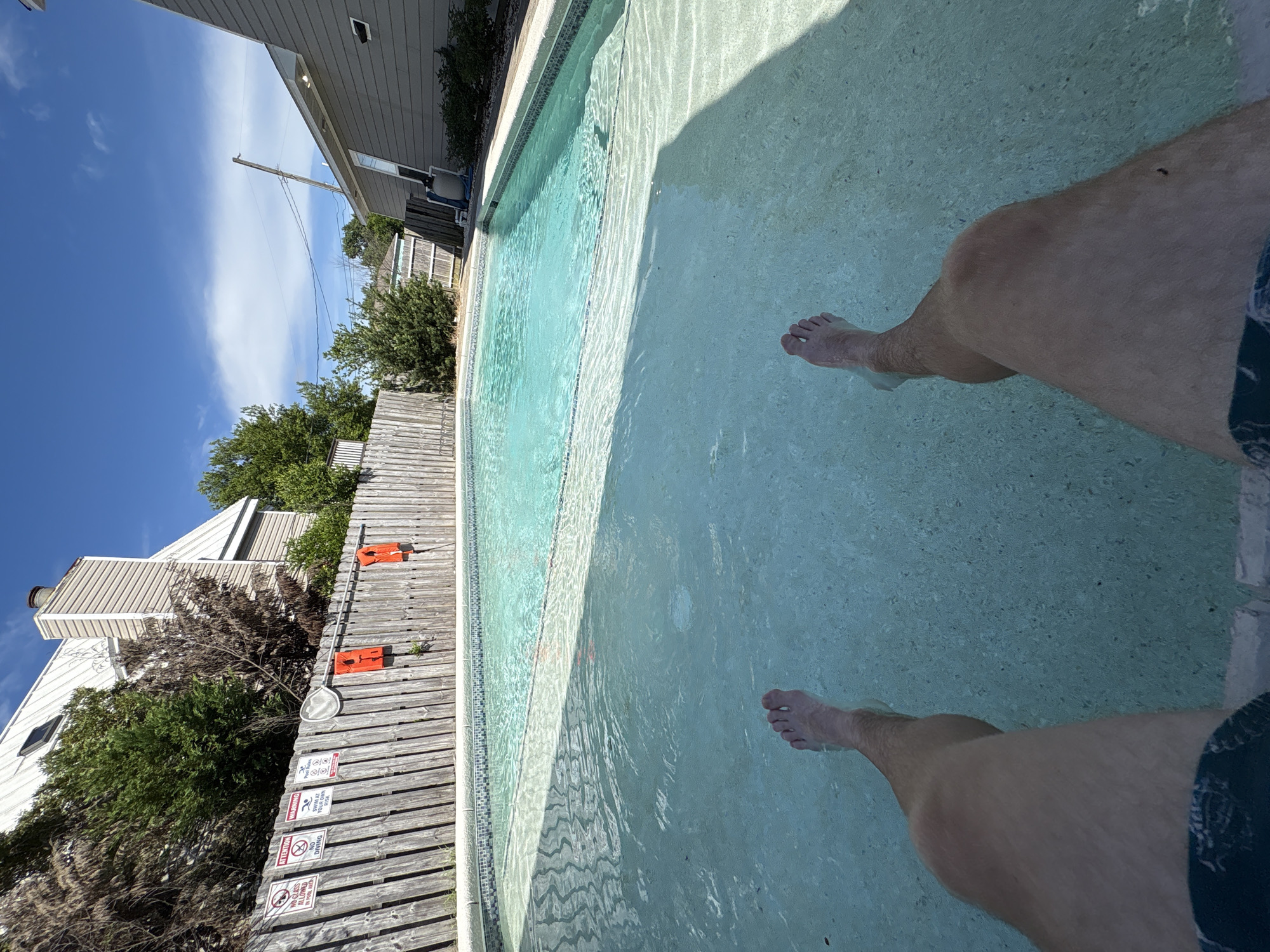 The image shows a first-person perspective of a person's legs submerged in a swimming pool. The pool water is clear with visible ripples and shadows on the bottom. The person is wearing dark patterned swim trunks. In the background, a wooden fence surrounds the pool area. Several warning signs are attached to the fence, including one that reads "NO GLASS IN POOL AREA" and another that says "ATTENTION NO DIVING." Two orange life vests are hanging on the fence. Beyond the fence, trees and a multi-story building with a light-colored metal roof are visible. The sky is blue with scattered white clouds. The scene appears to be a residential backyard in Panama City Beach, Florida, United States of America.