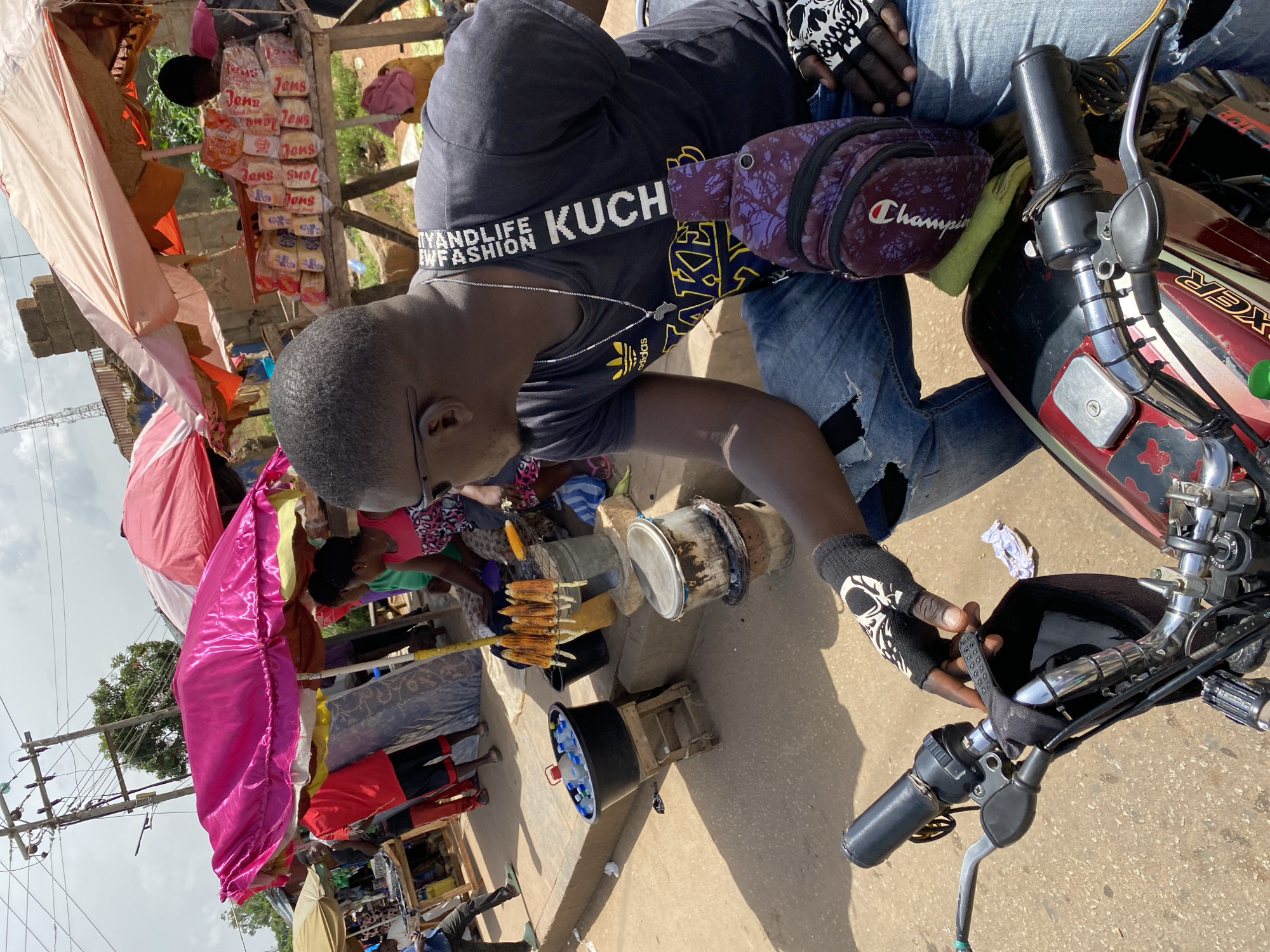 The image depicts a street scene in Bukuru, Nigeria. In the foreground, a person wearing a dark t-shirt and ripped denim jeans is seated on a motorcycle. They have on fingerless gloves with a skull pattern and a silver chain with an Africa pendant. A purple fanny pack with a "Champion" logo is slung across their chest. The motorcycle's handlebars and part of its body are visible.
Behind the person, a market stall is set up. Various items are displayed, including what appear to be packaged snacks or bread stacked on shelves. There is also a pink umbrella or canopy, and several people are gathered around the stall, engaged in what seems to be a transaction or browsing. In the mid-ground, a cylindrical object, possibly a stove or a smoker, is visible, along with a large black bucket containing bottles of water. A cluster of grilled corn on skewers is also present, suggesting food being sold.
The scene appears to be outdoors during the daytime, with natural light casting shadows on the ground. The presence of a market stall with goods and people interacting suggests a typical market day. In the background, there are some structures and vegetation, indicating a semi-urban or rural setting. Visible text includes "KUCH" and "IYANDLIFE EW FASHION" on the strap of the fanny pack and what looks like "Champion" on the fanny pack itself. The overall atmosphere seems active and transactional.