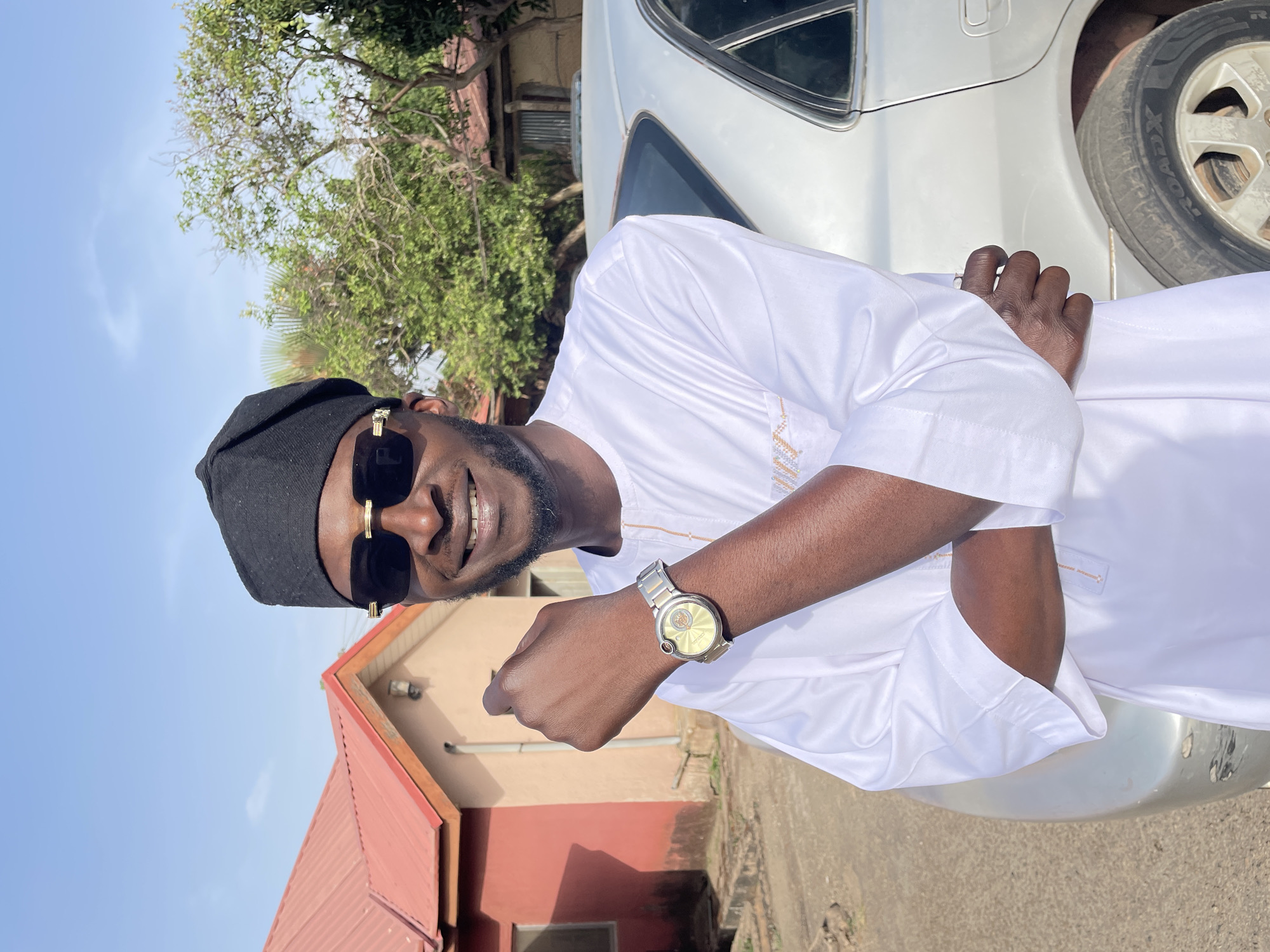 The image shows a smiling man wearing a black cap and sunglasses, standing in front of a silver car. He is wearing a white shirt with gold detailing and a silver watch. A building with a red roof is visible in the background, along with a tree and what appears to be a paved road. The location appears to be outside and the sky is blue.
