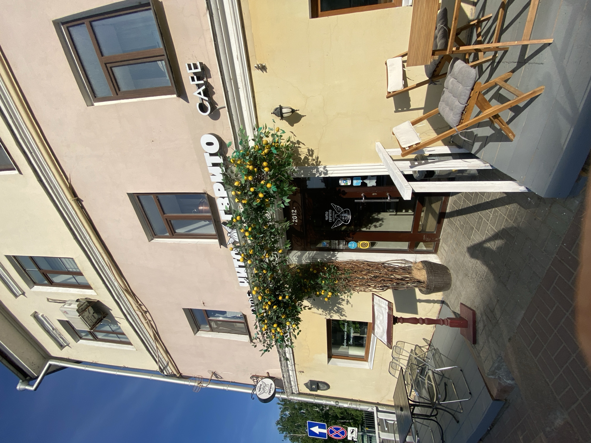 A cafe called "Чиto Твиrito Cafe" is located in Kazan, Russia. It is daytime. The cafe exterior is composed of beige and yellow painted walls. The cafe's name is displayed in white lettering on the building's facade. There are windows on the building. Outside, there are tables and chairs for outdoor seating. A lemon tree decoration adorns the entrance. Street signs are visible nearby. The sky is clear and blue. A menu stand is positioned near the entrance.