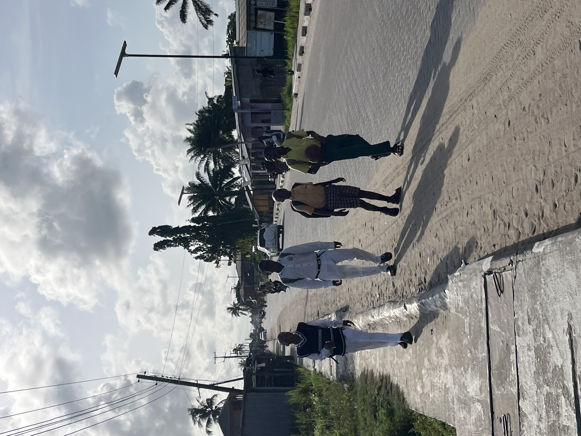 The image depicts an outdoor scene in Badagry, Nigeria. Four individuals are walking along a street. Two are wearing white and black uniforms, one is in a white uniform, and another in a yellow shirt. The street is lined with buildings, vegetation, and utility poles. The sky is cloudy. The street has tire tracks on it.