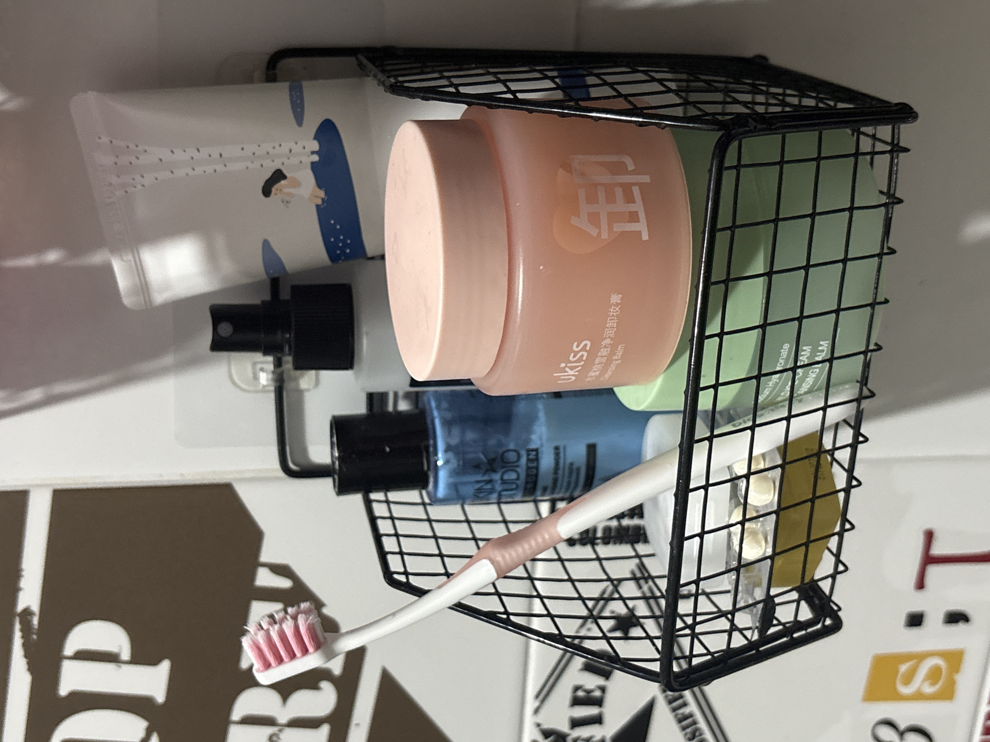 In the city of บ้านกาญจน์กนก 23, Thailand, a black wire basket is affixed to a wall, holding various personal care items. A pink and white toothbrush rests on the edge of the basket, pointing upwards and to the left. Inside the basket, a clear plastic container with yellow circular pills is visible at the bottom. Above this, a blue bottle with a black cap, labeled "KIN STUDIO", is partially obscured. To its right, a white tube with a minimalist illustration of a person sitting on a blue surface stands upright. In front of the blue bottle and the white tube, a round, light pink jar with a matching lid, labeled "ukiss" and featuring Chinese characters, is prominently displayed. A light green jar with white text, partially visible, sits behind the pink jar. The background consists of a tiled wall with some brown and white lettering.
