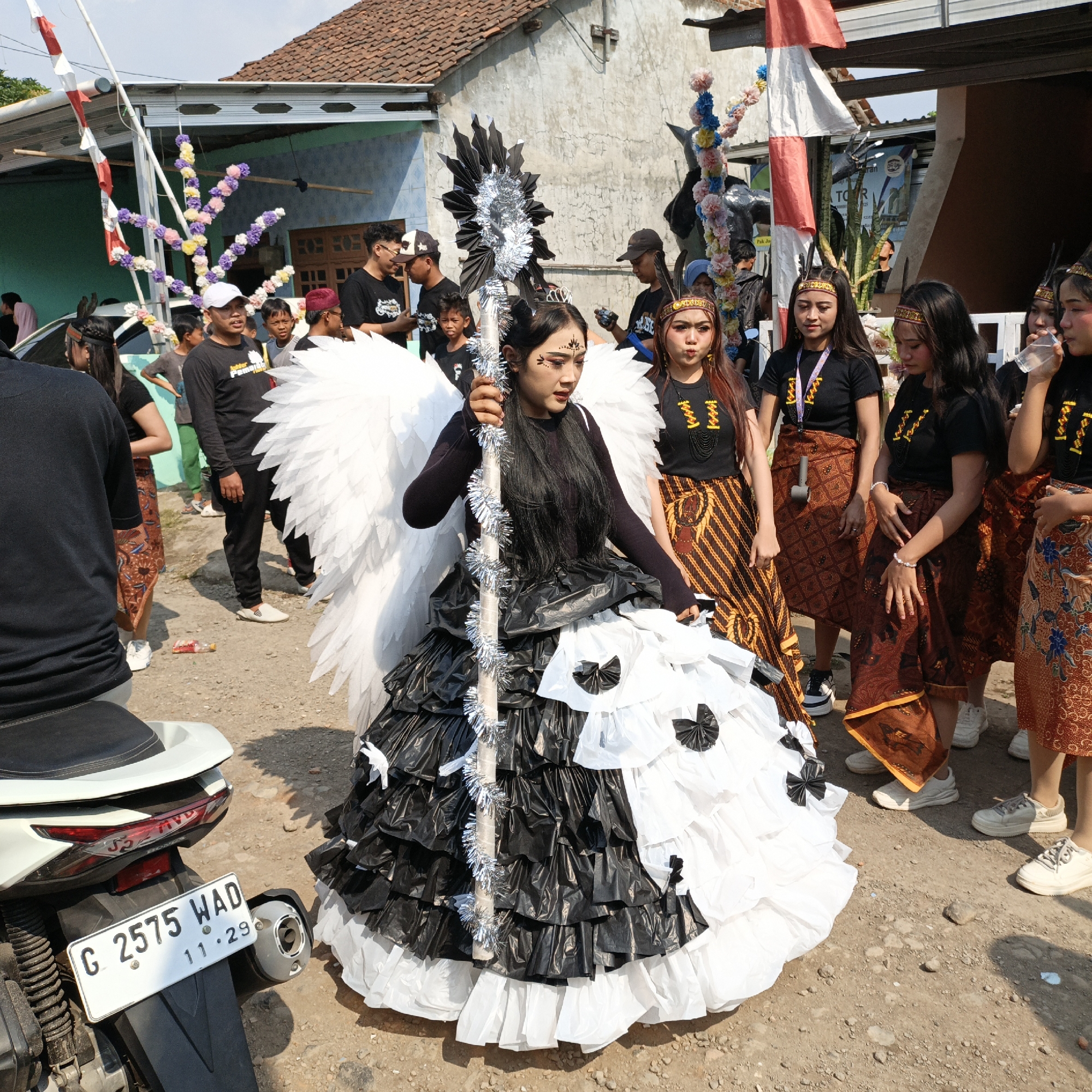 In Tanahbaya, Indonesia, a young woman stands dressed in an elaborate costume resembling an angel, adorned with large white wings and a layered black and white skirt made of plastic material. She holds a staff decorated with silver tinsel. Several other young women, wearing matching black tops and patterned sarongs, stand to her right, some holding drinks. In the background, a group of men and children are gathered, with a building and a motorcycle visible. The scene appears to be outdoors, possibly during a local event or parade, with a flag pole featuring red and white banners in the upper left of the frame.