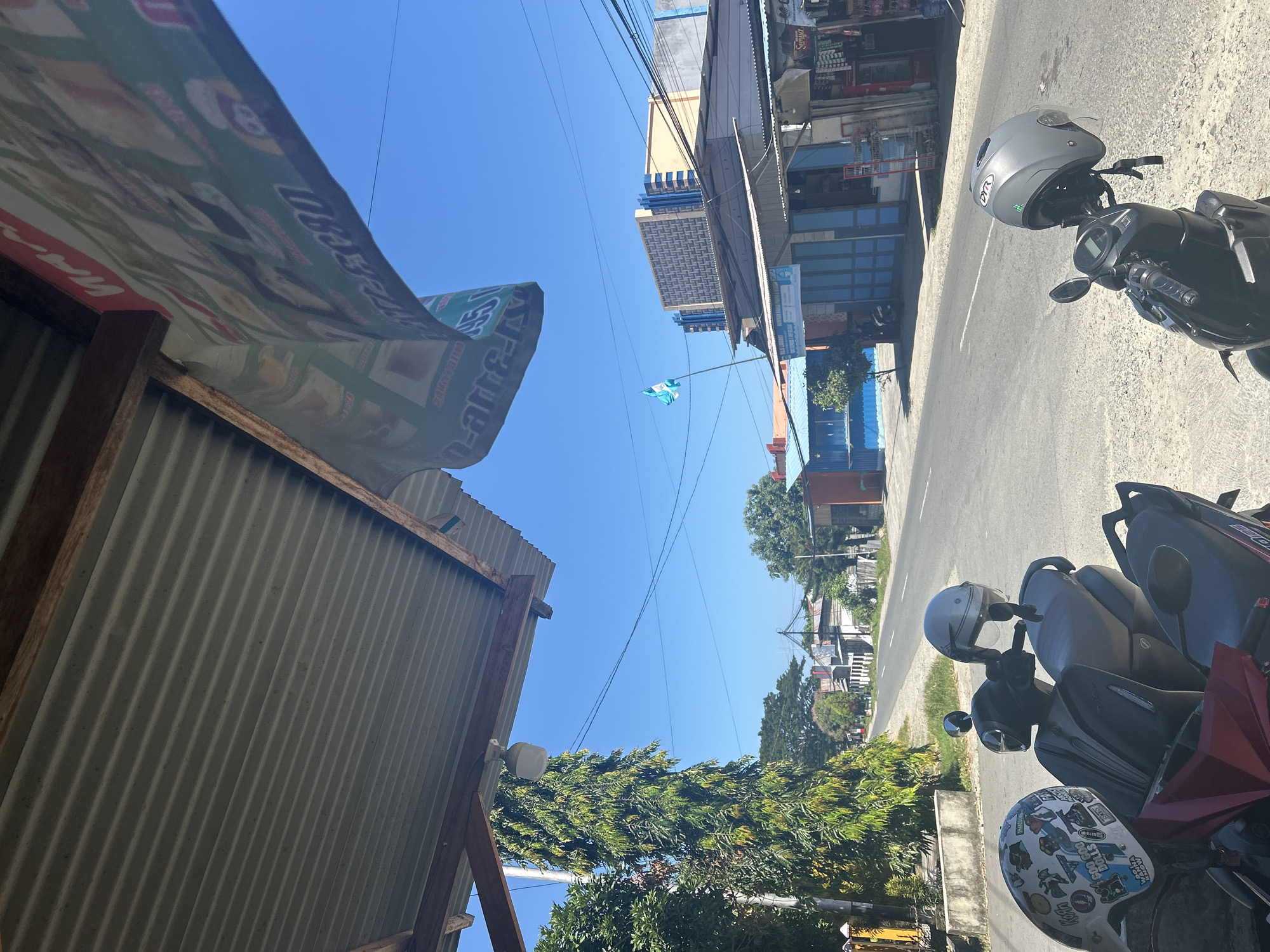 The image captures a street scene on a sunny day in Mandaong, Indonesia. In the foreground, two scooters are parked, each with a helmet attached. The scooter on the left has a white helmet adorned with various stickers, including cartoon characters and text. The scooter on the right has a grey helmet with a reflective visor. Both scooters are parked on a gravelly roadside.
Behind the scooters, a road stretches into the distance, lined with buildings and trees. The buildings appear to be shops and businesses, with awnings and signs visible. One prominent building in the mid-ground has a blue facade and blue folding doors. Further down the street, a tall building with a distinctive geometric pattern on its upper floors is visible. A flag with a light blue and white design flutters on a pole in front of this building.
Overhead, a corrugated metal roof extends from a structure on the left, casting a shadow. A banner with colorful images and text hangs from this roof structure, partially obscuring the bright blue sky. A street lamp is attached to a pole beneath the roof on the left. The overall environment suggests a typical urban or semi-urban street in Indonesia, with clear weather and bright sunlight.