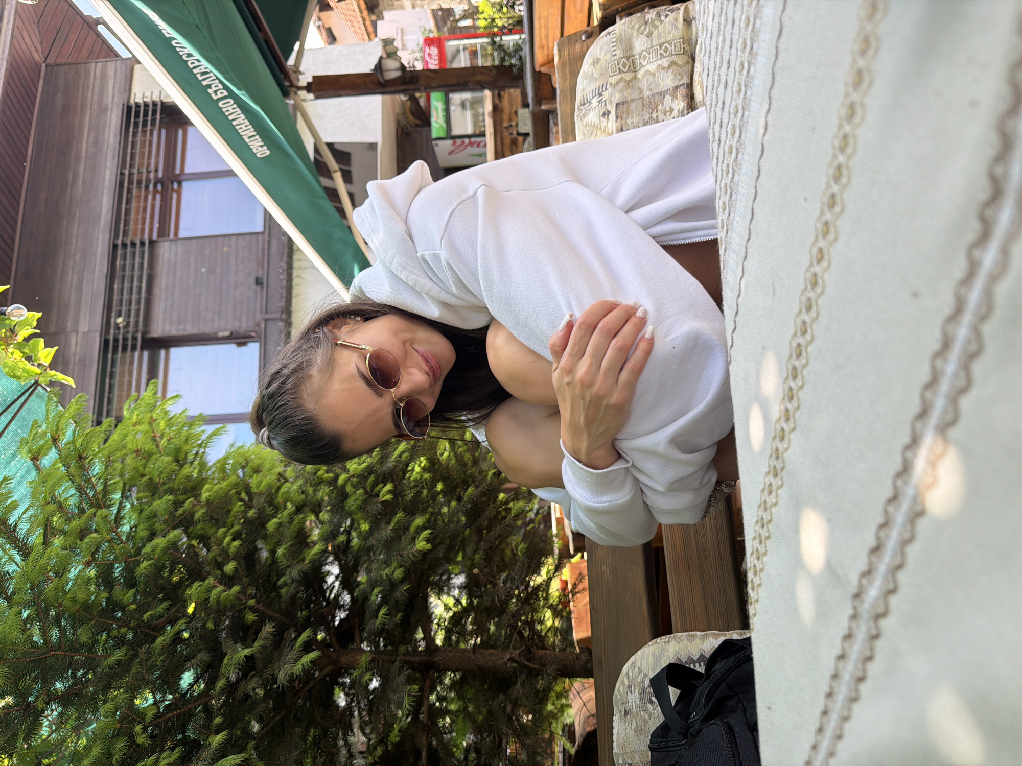 An outdoor scene captures a young woman seated on a wooden bench at a cafe or restaurant patio in an unknown city in Bulgaria. The woman, with dark hair pulled back, wears round, gold-rimmed sunglasses and a white hooded sweatshirt, smiling subtly while hugging her knees. Her fingernails are painted white. In the foreground, a light-colored tablecloth with two patterned brown and gold stripes is visible, illuminated by dappled sunlight. To the left, a black bag rests on a patterned cushion atop a wooden chair. The background features a large green coniferous tree on the left. Behind the woman, a two-story building with dark wooden siding and windows is visible, partially obscured by a green awning displaying white Cyrillic text, which translates to "ORIGINAL BULGARIAN..." (the full text is not visible, but implies "BEER" or similar). Further to the right in the background, a red Coca-Cola branded refrigerator and a white refrigerator are situated against another building. The setting is bright and sunny.