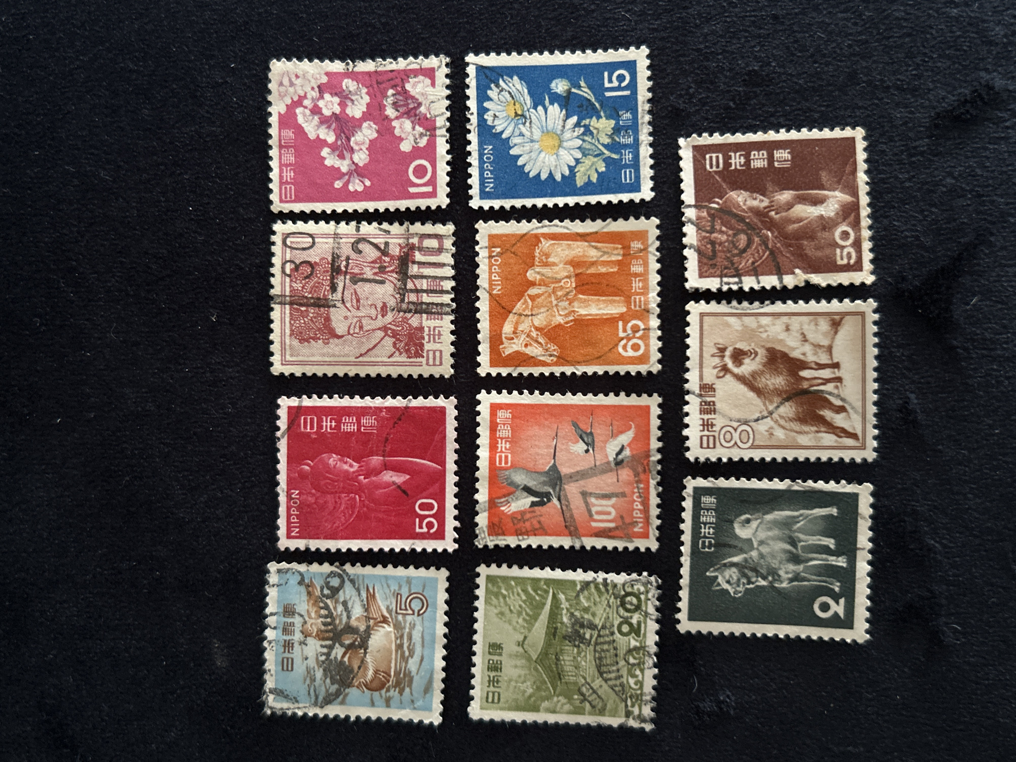 The image displays a collection of twelve postage stamps arranged in three rows and four columns against a dark, textured background. The stamps feature a variety of designs, including animals, floral motifs, and human figures, with Japanese characters present on most.
The top row, from left to right, shows a stamp with a bird and water, a stamp with a red background and a figure, a stamp with a pink background and a figure, and a stamp with a pink background and cherry blossoms. The second row, from left to right, features a stamp with a green background and a building, a stamp with an orange background and birds in flight, a stamp with an orange background and a horse, and a stamp with a blue background and daisies. The bottom row, from left to right, contains a stamp with a dark green background and a dog, a stamp with a brown background and a goat, and a stamp with a brown background and a seated figure.
The stamps appear to be in good condition, with visible perforations along their edges. The image is well-lit, allowing for clear observation of the details on each stamp. The dark background helps to make the colorful stamps stand out. The overall impression is that of a carefully arranged display of collectible items.