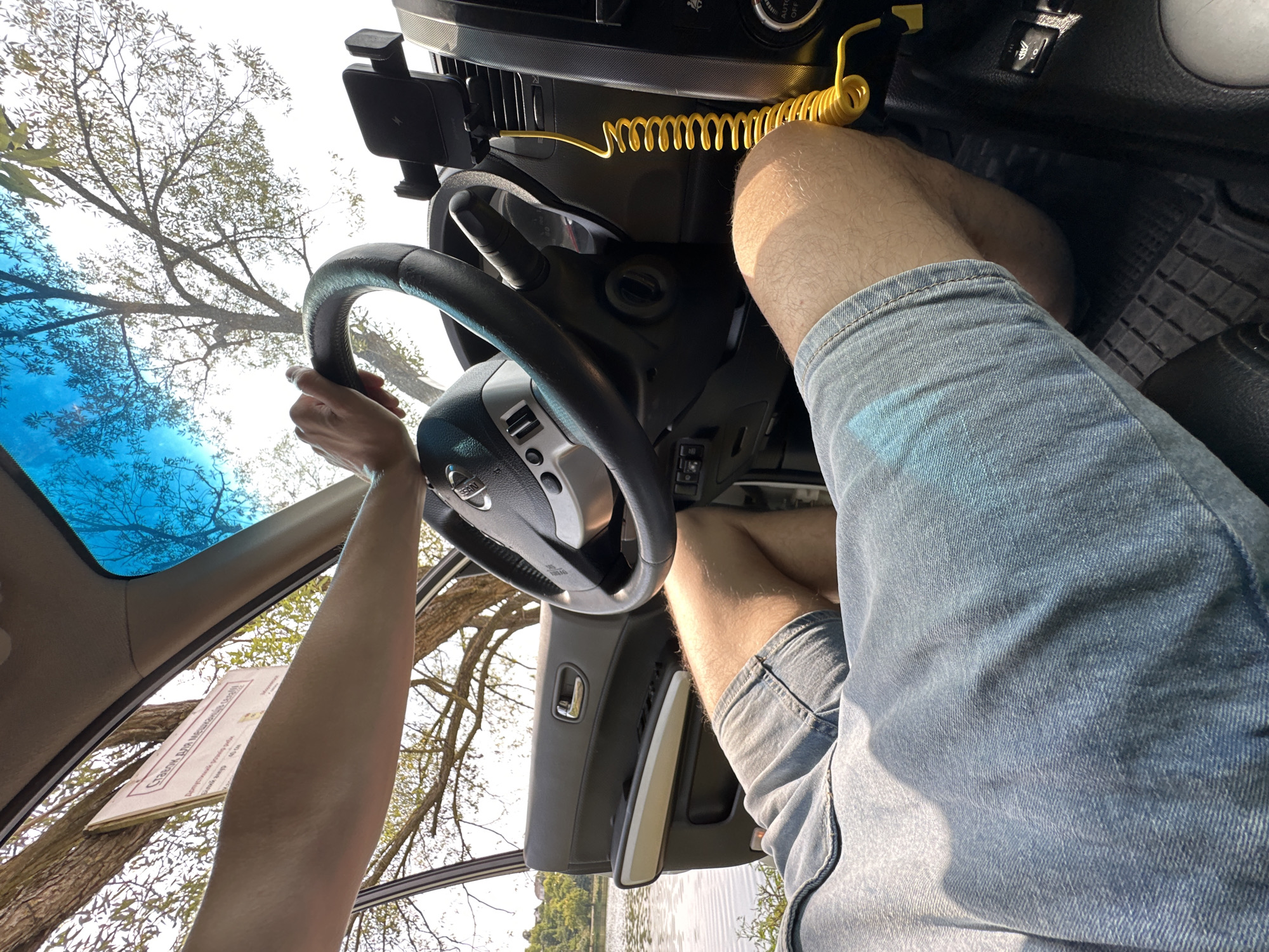 A first-person perspective shot from inside a car shows a person driving near a body of water. The driver's hands are on the steering wheel, and their legs are visible in denim shorts. The car's interior, including the dashboard, steering wheel, and a phone mount with a yellow cord, is in focus. Outside the car, a tree with green leaves and a blue sky are visible. The car appears to be parked near the shore of a lake or river, with trees and buildings in the background, suggesting a natural or semi-rural setting. The image captures a moment of leisure or travel, with the driver enjoying the scenery. The specific location, Makiv, Ukraine, is confirmed by a sign visible on the car's windshield which translates to "Rules for Fishing" in Ukrainian, a common regulation sign found in Ukrainian fishing areas.