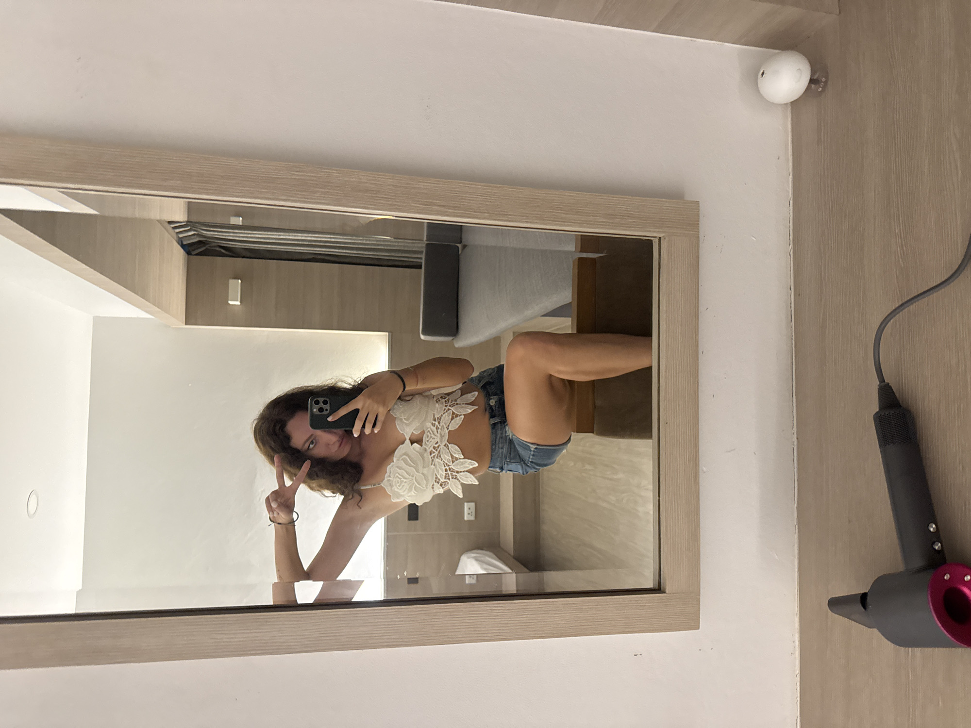 A young woman with long, curly brown hair is taking a mirror selfie in what appears to be a hotel room in Koh Samui, Thailand. She is wearing a white lace bralette with floral appliques and denim shorts. Her left arm is raised, and she is making a peace sign with her fingers. She is holding a smartphone in her right hand, which is angled to capture her reflection.
In the reflection of the mirror, a portion of a bed is visible, along with a bedside table and wall-mounted electrical outlets. The room appears to have light-colored wood paneling on the walls and a wooden floor. The overall aesthetic suggests a modern, minimalist hotel setting.
In the foreground of the image, on a wooden surface, a dark gray and pink Dyson hairdryer is positioned horizontally. To the right of the hairdryer, a small, white, round object, possibly a diffuser or air freshener, sits on the floor. The wall to the right of the mirror is a pale, neutral color.