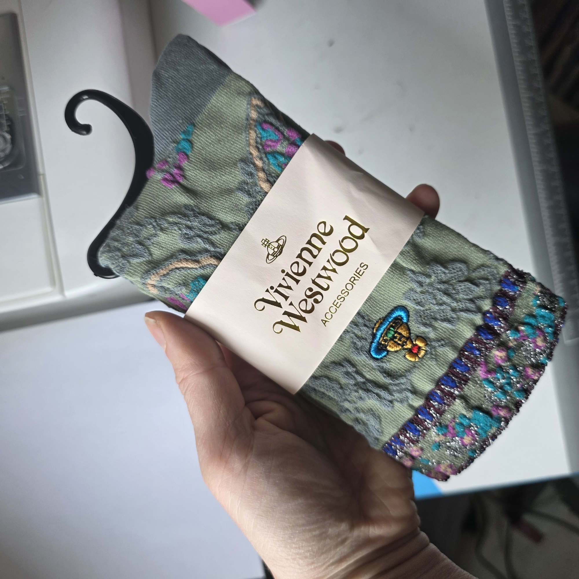 A hand holds a pair of Vivienne Westwood socks on a black plastic hanger. The socks are pale green with floral-like embroidered patterns and a stripe of colorful squares at the cuff. The Vivienne Westwood logo is embroidered on the sock. The socks are wrapped in a white paper band with "Vivienne Westwood Accessories" printed in gold. The background is a white surface with a blurry image of a box on the left and a straight edge on the right.
