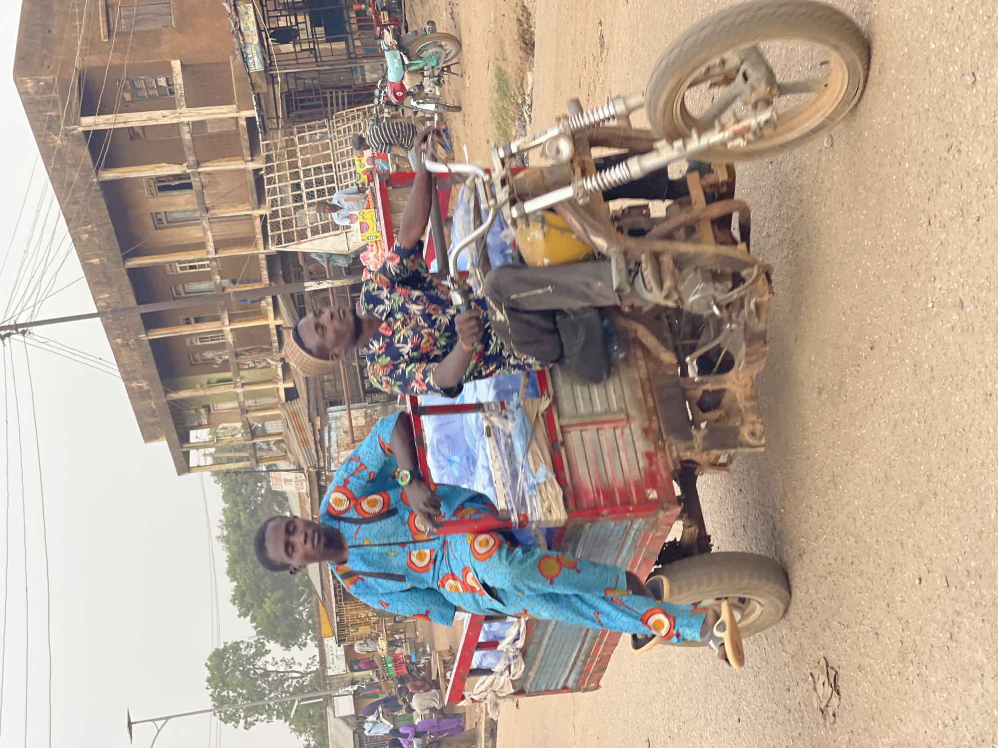This photograph captures a vibrant street scene in Chikaji, Nigeria, featuring two men on a motorized tricycle, commonly known as a Keke Marwa or Napep. The image is taken from a slightly elevated perspective, looking down on the vehicle and the surrounding environment.
In the foreground, a man is driving the tricycle. He wears a dark, short-sleeved shirt with a bright floral pattern and a light-colored cap. His hands are on the handlebars, and a yellow container, possibly for fuel, is visible near his feet. Seated in the cargo bed behind him is another man, dressed in a striking blue two-piece outfit adorned with orange and white circular patterns. He stands upright in the back, facing the camera.
The tricycle itself appears functional but rugged, with visible wear and rust on its metallic parts. The cargo bed is red with blue tarp-like material lining its interior. The vehicle is on an unpaved or dirt road, characteristic of many local roads.
In the background, there are multi-story buildings, some appearing residential or commercial, with basic construction and exposed concrete elements. Electrical wires crisscross overhead. Other individuals and a parked motorcycle are faintly visible further down the street, adding to the bustling atmosphere of the setting. Trees and sparse vegetation dot the landscape. The sky is overcast, suggesting daytime with diffused light, possibly late morning or afternoon. The overall scene conveys a typical daily moment in a Nigerian community.
Visible text on signs in the background is blurry, but one sign appears to read "Menu &" and another might have initials like "C B". The emotions of the visible individuals appear neutral.