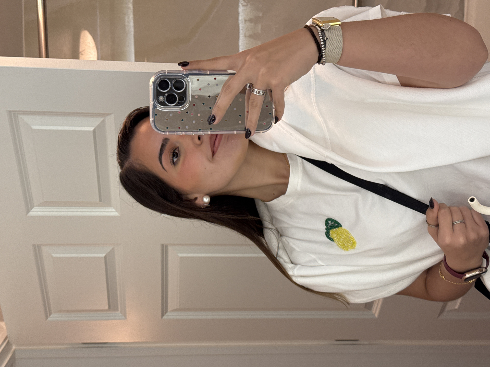 A woman is taking a selfie in front of a white door. She is wearing a white T-shirt with a yellow design, a silver watch, and several rings. The background is blurry, but it appears to be a bathroom in a residential area, likely in San Antonio, United States of America.