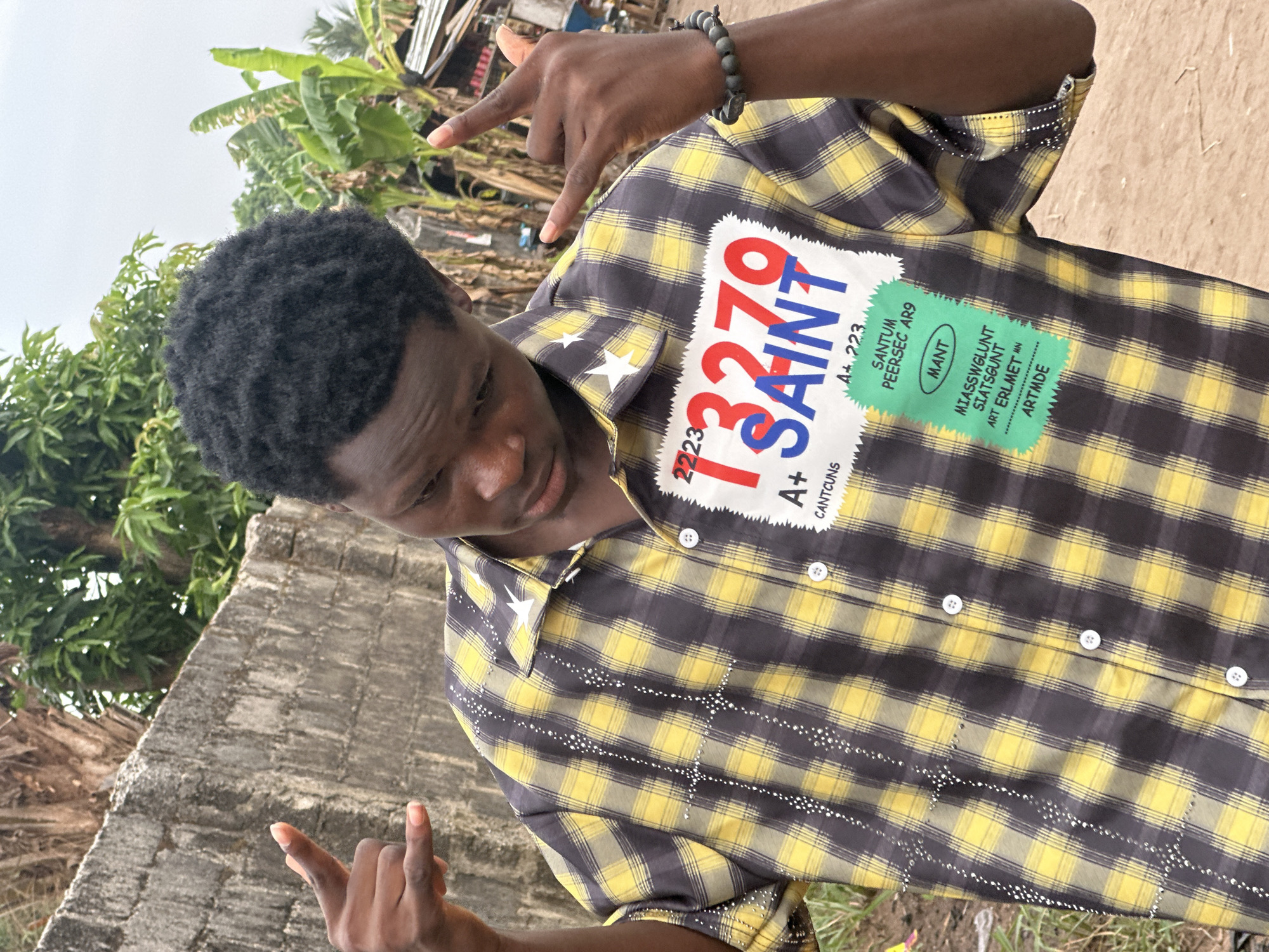 A young dark-skinned male is captured in a half-body shot, posing outdoors in Ihiagwa, Nigeria. He wears a short-sleeved, collared shirt featuring a black and yellow plaid pattern with white buttons down the front. The shirt is adorned with white star patches on the collar tips and small, reflective studs arranged in vertical and horizontal lines across the upper left front, forming a cross-like design. Similar studs are visible on the sleeve cuffs. A large, multi-text white and green patch is prominently displayed on the upper right front of his shirt, featuring red numbers "13279" and blue text "SAINT," along with other alphanumeric details such as "2223," "A+ CANTCUNS," and "SANTUM PEERSEC AR9." A dark, beaded bracelet is on his left wrist. He is performing a "rock on" hand gesture with both hands, extending his index and pinky fingers while curling his middle and ring fingers. His gaze is directed downwards and to his left. The background features a grey concrete block wall on the left, and dense tropical vegetation, including banana plants, along with indistinct structures possibly made of corrugated metal, on the right. An overcast sky is visible above the vegetation. The ground appears to be dirt or sparsely vegetated.