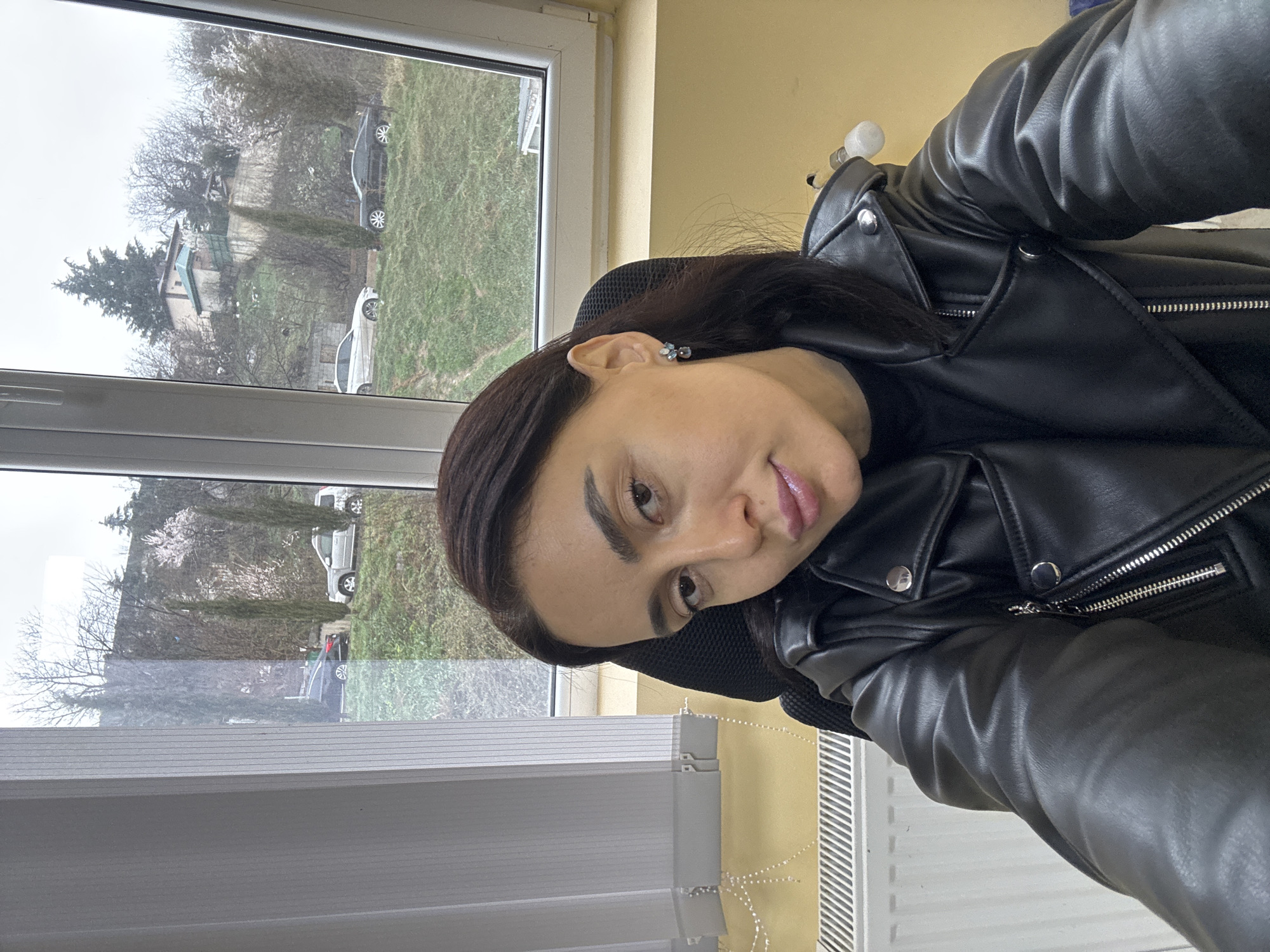 A woman with dark brown hair and fair skin is depicted taking a selfie indoors, seated in a black mesh office chair. She wears a black mock-neck top and a black leather-look motorcycle jacket with silver zippers and snap buttons. A silver-colored earring with light blue/grey stones is visible on her right ear. The room features pale yellow walls and a white vertical radiator along the lower left. A large window with a white frame occupies the background, partially covered by white vertical blinds on the left.
Through the window, a hilly, semi-urban landscape is visible under an overcast sky, consistent with the city of Tbilisi, Georgia. The hillside is covered with green grass and features various trees, including some deciduous trees with sparse foliage and one with light-colored blossoms, alongside taller evergreen trees. Several vehicles are parked on the hillside: a dark sedan, a silver SUV, and parts of other sedans (one dark, one light-colored). In the upper right background, a small, light-colored house with a green roof is situated on the slope amidst trees.