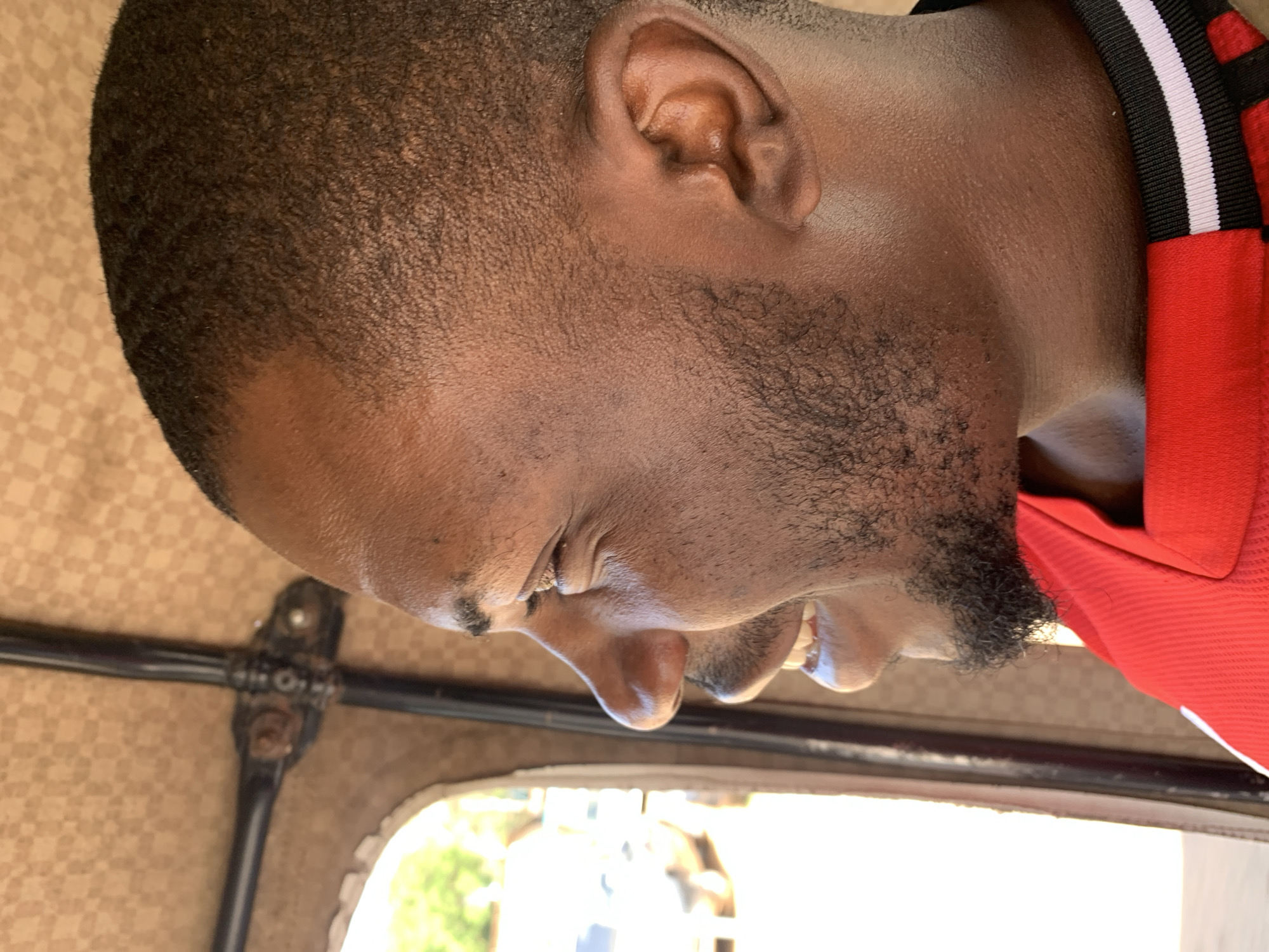 The image, oriented on its side, captures a close-up side profile of a man with dark skin, short, closely-cropped dark hair, and a neatly trimmed beard or goatee. He is wearing a red collared shirt featuring black and white stripes on the collar. His right ear is prominent, and his face is angled downwards, suggesting he is looking out. His right eye appears to be squinting slightly, possibly due to bright ambient light.
The setting places the man inside a vehicle. The background reveals a patterned fabric ceiling, tan and brown with a diamond grid design, and blurred metal structures, likely part of the vehicle's frame. Through an open window or side, a brightly lit outdoor scene with some distant greenery and vague shapes is visible. The intense light suggests it is daytime. This type of patterned interior and metalwork is common in motorized tricycles, often referred to as 'Keke Napep' in Nigeria. The man appears to be observing the outside environment from within the vehicle. The scene is situated in Rafin Par, Nigeria. No visible text is present in the image.