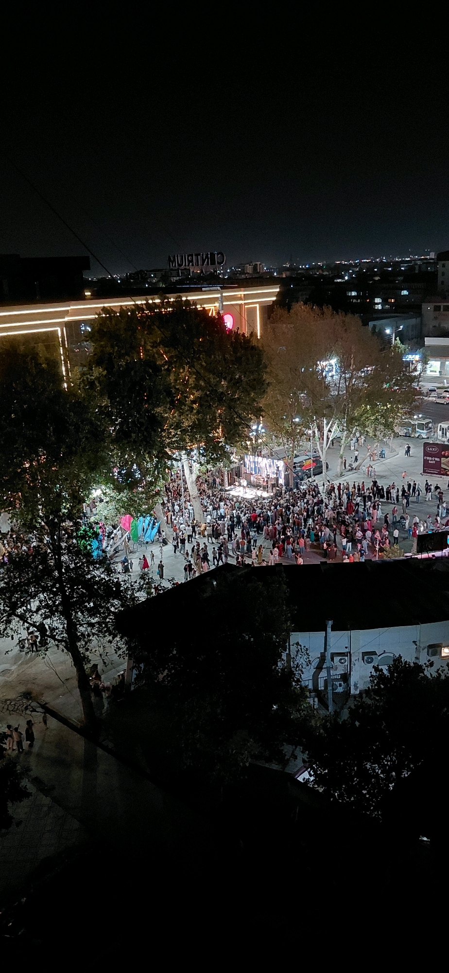 The image captures a night scene in the city of Samarkand, Uzbekistan, likely an outdoor public gathering space or event. In the background, a large building is illuminated with horizontal strip lights and a prominent sign that reads "CENTRUM" in large, illuminated letters. Several trees with dense foliage are scattered throughout the scene, some lit from below, casting shadows.
The focal point of the image is a densely populated area in the mid-ground. A stage or platform is set up with bright lights, suggesting a performance or event is taking place. A large crowd of people, appearing to be of various ages and dressed in casual attire, have gathered around the stage and are spread out into the surrounding area. Some individuals are standing, while others appear to be walking. The atmosphere suggests a lively event, possibly a concert, festival, or public celebration.
In the foreground, a dark, silhouetted building with some visible details, such as air conditioning units and windows, occupies the lower portion of the image, partially obscuring the view of the scene above. The overall lighting is dim, characteristic of nighttime, with artificial lights from the buildings, stage, and streetlights illuminating the activity. The image is taken from a high vantage point, looking down onto the scene.
