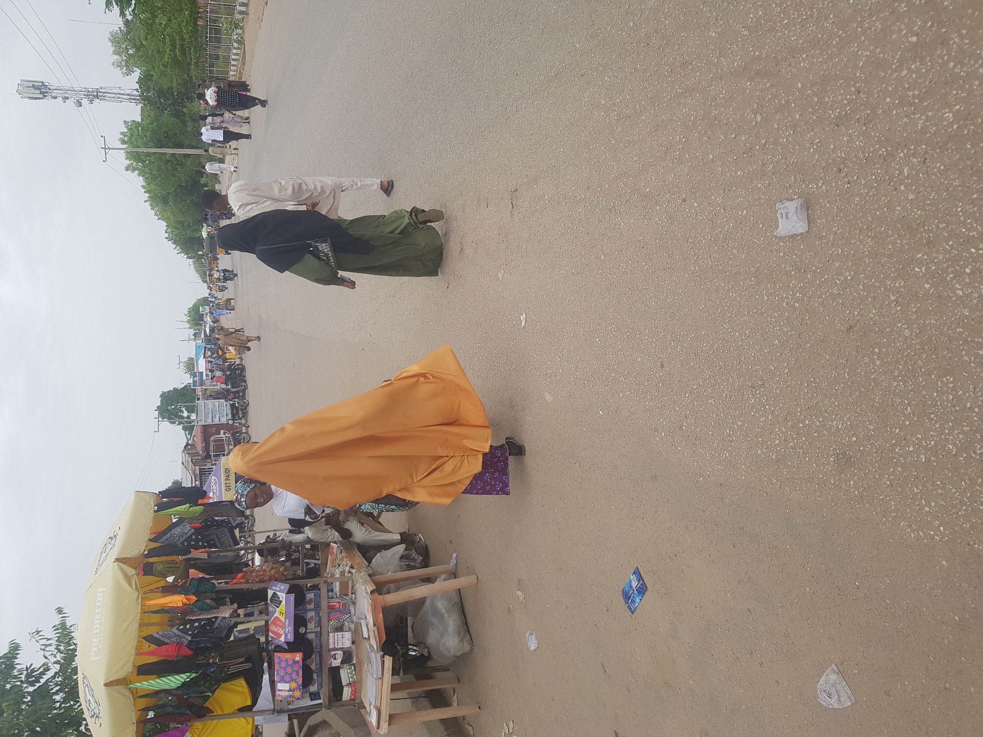 The scene is an outdoor street in Dabawa, Nigeria. The ground appears to be a paved road with some debris including a torn wrapper and paper trash. An outdoor vendor is selling items under an umbrella. One person is crouched behind the vendor's stand wearing a peach-colored dress. There are several people walking in the street. One person is wearing a black covering and a green dress. Another man is wearing a white robe. Several other men can be seen in the background. In the distance, a cell tower is visible.