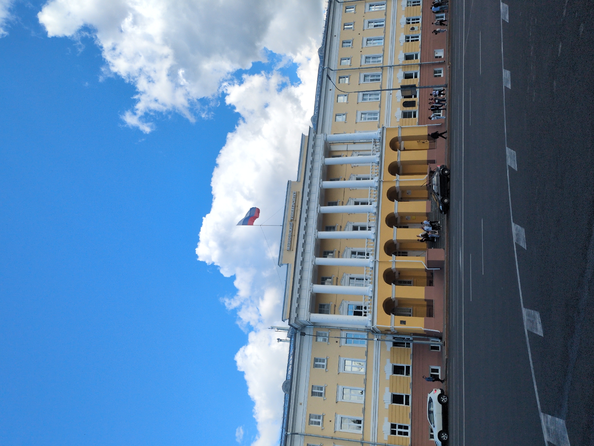 This is a high-angle, vertical photograph of a yellow building with white columns, situated along a street. The building appears to be a public institution, possibly governmental or educational, with multiple floors and many windows. A Russian flag is visible flying from a flagpole on the building's facade. Several people are seen walking on the sidewalk in front of the building and seated on steps. A black car is parked near the entrance of the building, and a white car is parked further down the street. The street itself is paved and marked with white lines. The sky is a bright blue with large, fluffy white clouds, suggesting it is daytime with good weather. The overall scene is one of an urban environment on a clear day. The text "МИНИСТЕРСТВО ОБРАЗОВАНИЯ" (Ministry of Education) is visible on the building.