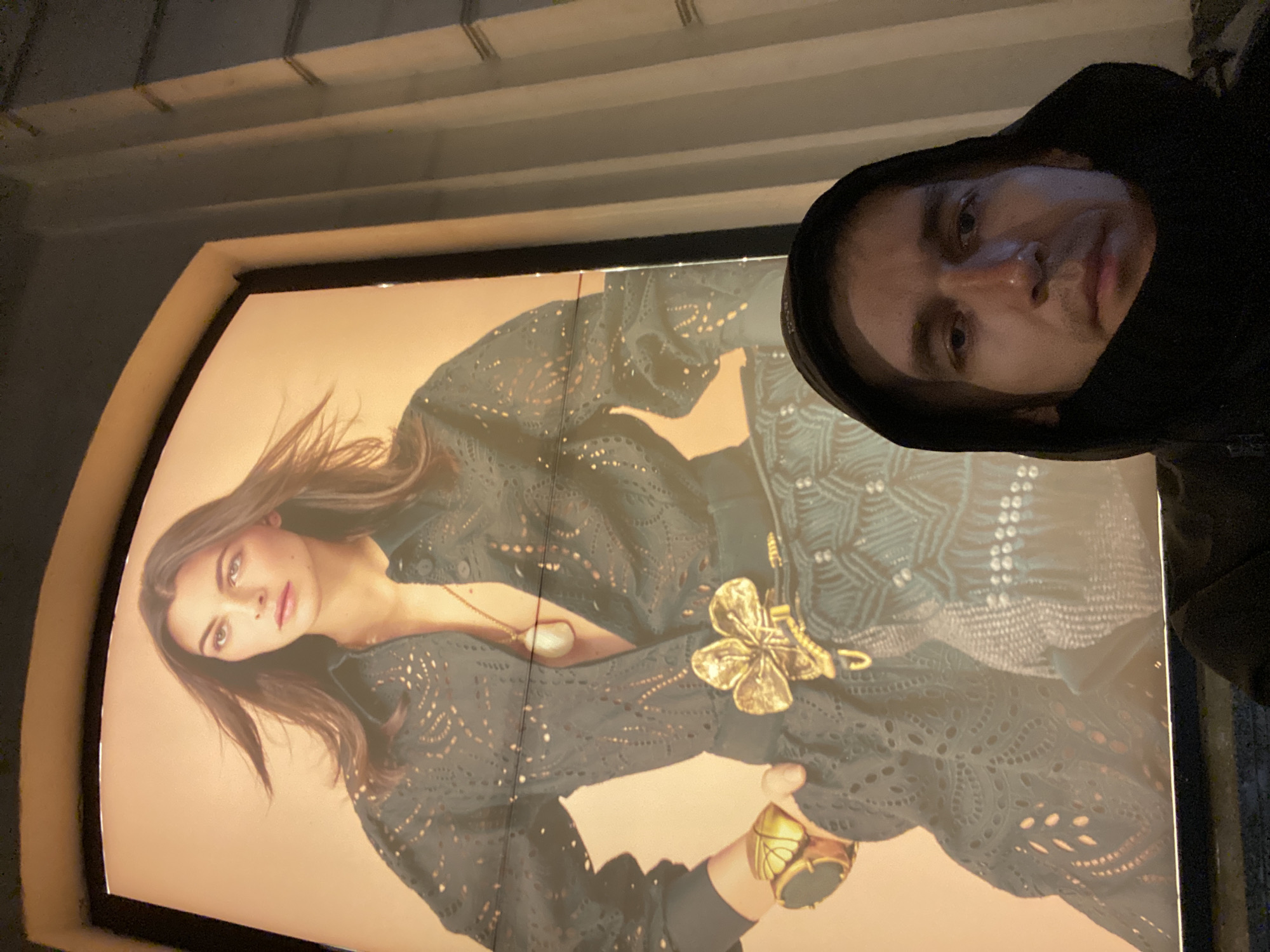 The image is a selfie taken by a person wearing a black hoodie, with a fashion advertisement poster in the background. The poster features a woman with long brown hair, wearing a dark, intricately patterned lace outfit. She is adorned with a large, ornate gold butterfly brooch and a chunky gold ring. The background of the poster is a soft peach color.
The selfie is taken from a slightly low angle, with the person's face partially visible in the upper right corner of the image, looking directly at the camera. The lighting in the scene is somewhat dim, suggesting it might be evening or an indoor setting. The person's expression is neutral.
The overall impression is casual, with the person capturing a moment perhaps while passing by the advertisement. There is no visible text in the image. The arched frame of the illuminated poster and the ceiling structure above suggest an indoor advertising display, possibly in a shopping mall or public space. The location context provided (Novokuybyshevsk, Russia) offers geographical information but cannot be inferred from the visual content of the image itself.