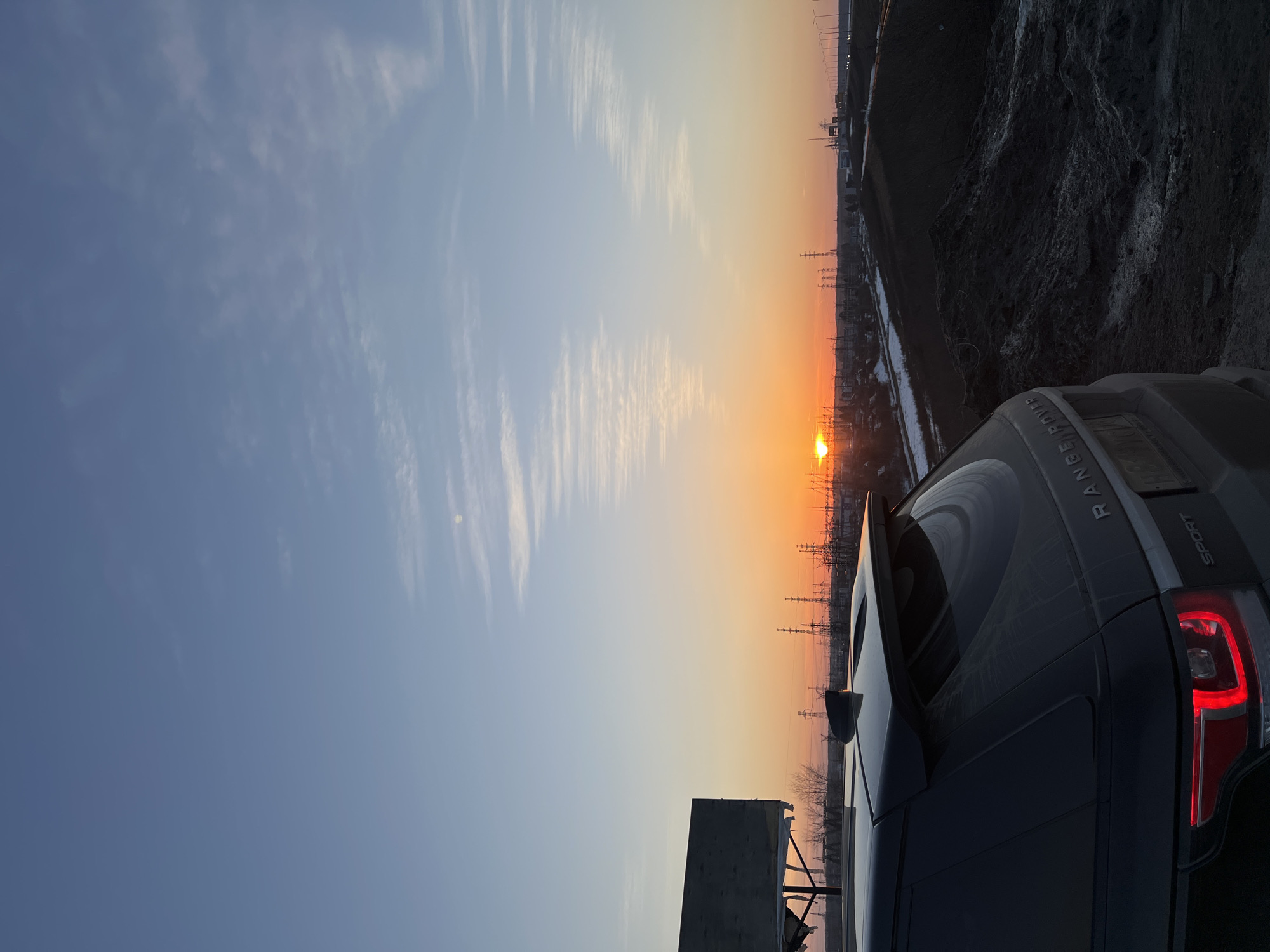 The image shows a sunset in what appears to be the city of Chaikovsky, Russia, evidenced by the presence of numerous high-voltage power lines, a feature commonly seen in industrial areas and near power plants. Part of a dark-colored Range Rover Sport is visible in the foreground, parked at an angle. The sky transitions from blue to orange, with the sun positioned on the horizon. Streaky cirrus clouds are visible in the upper portion of the sky. A building or structure partially obscured by other elements is situated in the lower-left. A snowy area is visible on a hill behind the powerlines.