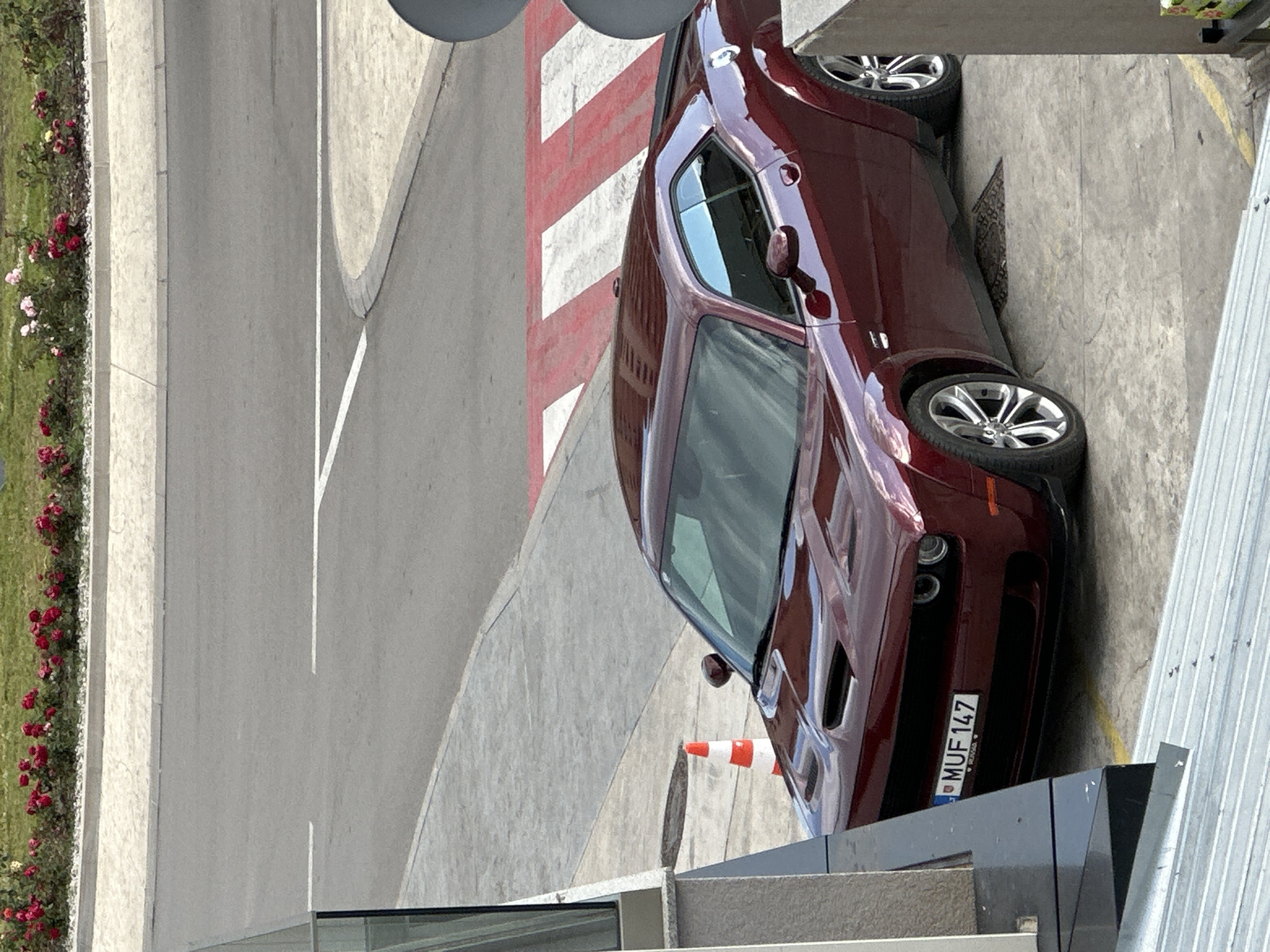 A maroon Dodge Challenger is parked near a building in Donji Pobori, Montenegro. The car's license plate reads "MUF 147." A traffic cone is visible near the front of the vehicle. To the left is a two-lane road with a flower bed alongside it. A crosswalk with red and white stripes is partially visible. The photograph appears to be taken from an elevated position.