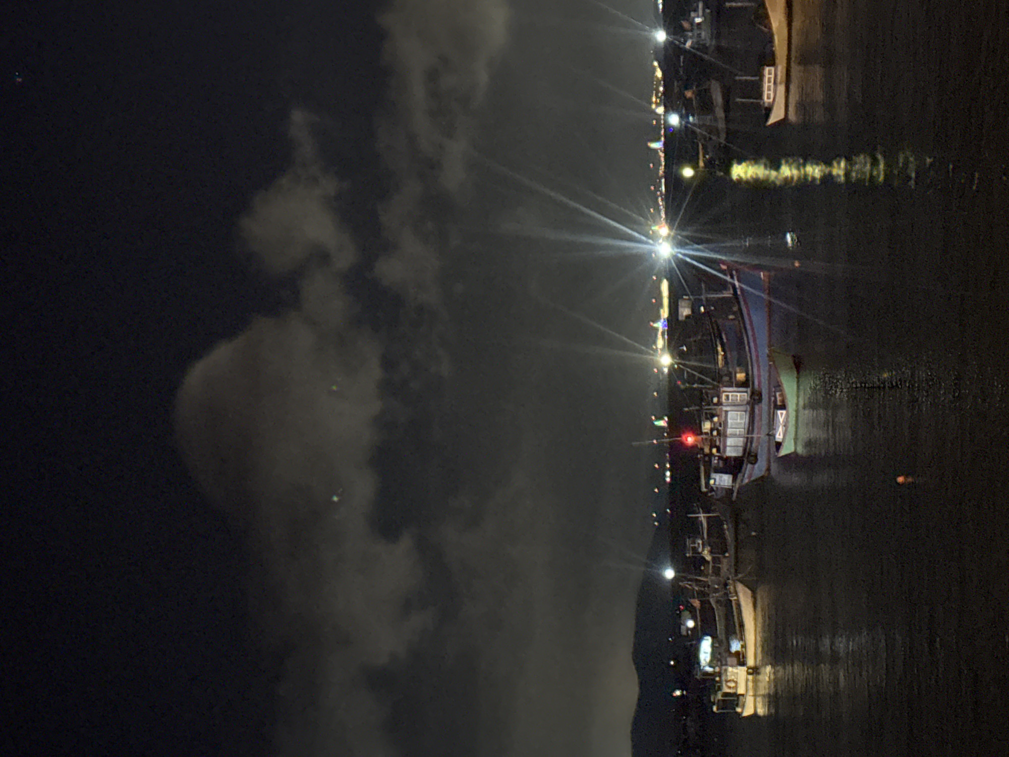 The image depicts a nighttime scene of a harbor in Bac Nha Trang Ward, Vietnam. Several fishing boats are anchored in the dark water, their lights reflecting on the surface. The sky above is filled with dark, cloudy formations, with distant city lights visible on the horizon. Some of the boats have their lights on, illuminating their decks and surroundings. The overall atmosphere is quiet and serene, characteristic of a harbor at night.