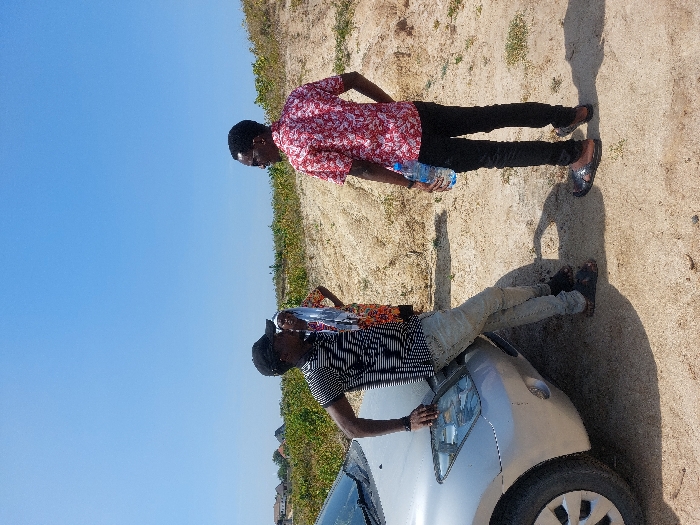 The scene depicts three individuals and a silver car on a dry, light-brown earthen surface under a clear blue sky in Kibaha, Tanzania.
On the left, a male wearing a black baseball cap, a black and white striped shirt, light-colored jeans, and dark sandals leans with his right hand on the front of a silver passenger vehicle. A dark wristband is visible on his left wrist. The vehicle's front bumper, headlight, and part of the driver's side wheel are visible.
To the right of the car, a second male stands with his back partially turned, facing the first individual and a third person. He wears a red and white patterned short-sleeve shirt, black trousers, and dark sandals. He holds a clear plastic water bottle in his left hand.
Between the two males, slightly in the background, a third individual, identifiable as female, stands with her hands on her hips. She wears a multi-colored patterned garment and a light-colored head covering.
The ground consists of dry soil with sparse green vegetation, particularly prominent to the right and in the mid-ground. In the far background on the left, several buildings with dark roofs are visible, indicating a nearby residential area. Strong sunlight is present, casting distinct shadows from the people and the car.