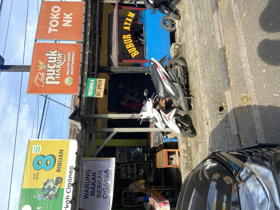 The image shows a street scene in Jatiluhur, Indonesia (latitude: -6.565, longitude: 107.434). A white scooter is parked on the sidewalk, angled to the right, next to a blue and red food cart with a yellow sign reading "Bubur Ayam" in Indonesian, meaning "chicken porridge." The cart is positioned against a building with a green sign that reads "Musholla WC Umum", indicating a place of worship with public restrooms. To the right of the food cart, there is a black motorbike parked on the road. Behind the scooter and food cart, there is a building with several signs. One sign reads "Warung Makan Berkah Ciganea", indicating a restaurant called "Ciganea". Another sign reads "8 Ribuan", likely referring to a price. The background includes a large sign in orange with a red and green logo that reads "Minuman Teh Pucuk Harum Rasa Teh Terbaiik ada di Pucuknya!" in Indonesian, which translates to "Drink Pucuk Harum Tea, The Best Tea Flavor is in Pucuk". There is also a sign to the right of the Pucuk Harum sign that reads "Toko NK". The image is taken from a low angle, showing the sidewalk and the street in front of the building. The sidewalk is made of concrete and cobblestone, and the street is paved. There is a shadow cast on the ground in the lower right corner of the image. The sky is partially visible in the upper right corner. A person is partially visible on the left side of the image, walking towards the street. The scene is midday and there is sunlight. The scene is typical of a street scene in Indonesia.