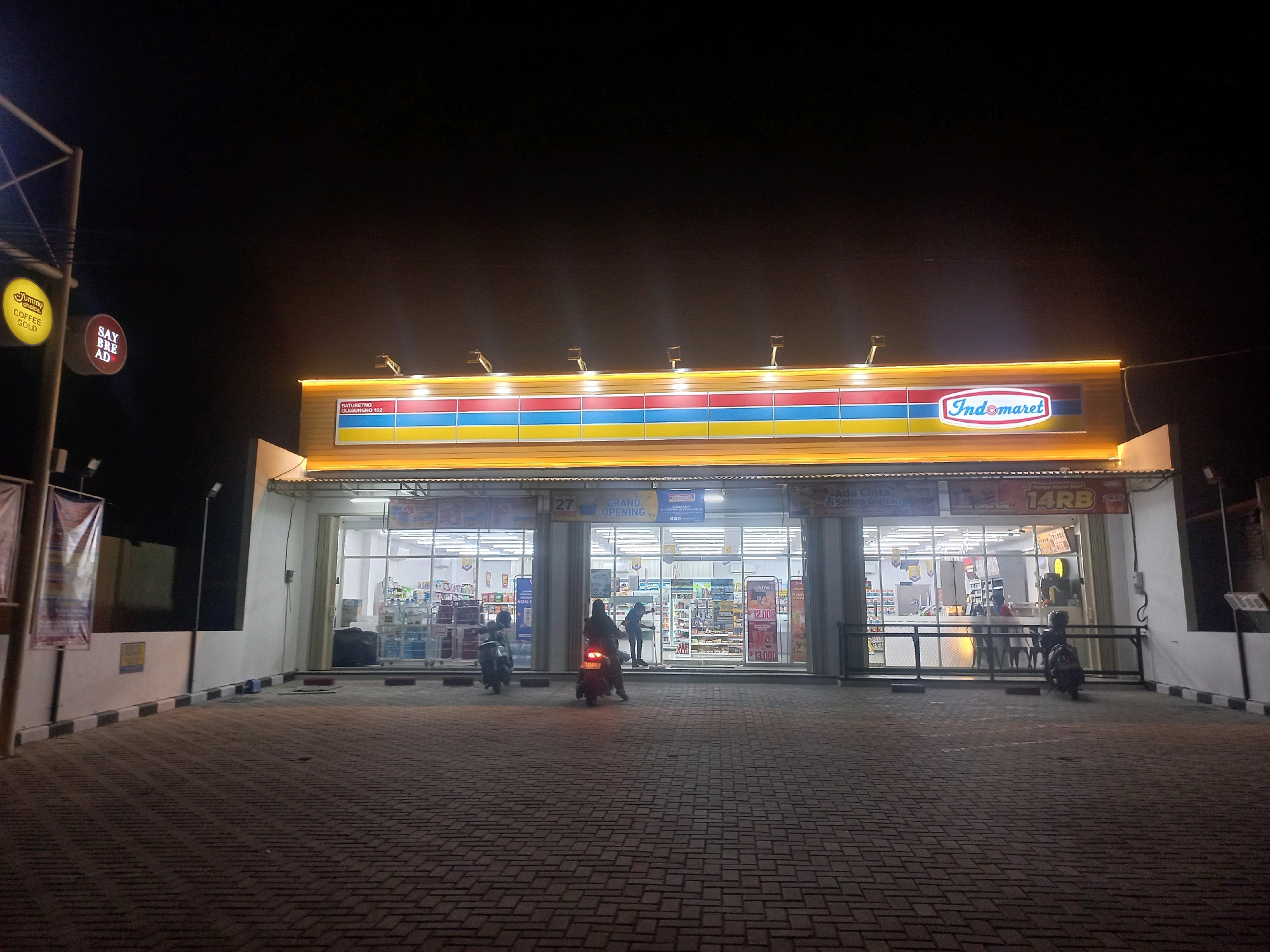 A night-time, eye-level, wide shot captures the exterior of an Indomaret convenience store in Baturetno, Indonesia. The store's illuminated facade features prominent signage, including the Indomaret logo and a horizontal banner with colored stripes (red, blue, yellow). The store's interior is visible through large glass windows, displaying shelves stocked with merchandise and illuminated by fluorescent lighting. In front of the store, a paved area is illuminated by the store's lights and streetlights. Three motorcycles are present: one is parked in the foreground to the right, another is partially visible in the middle ground to the left, and a third is in the middle ground, carrying two individuals. One person stands near the entrance, and another sits on a motorcycle. Two vertical signs are attached to a pole on the left side of the frame, one circular yellow sign with the text "Yummy Coffee Gold" and a circular red sign with white text. A white and black striped curb runs along the edge of the paved area.
