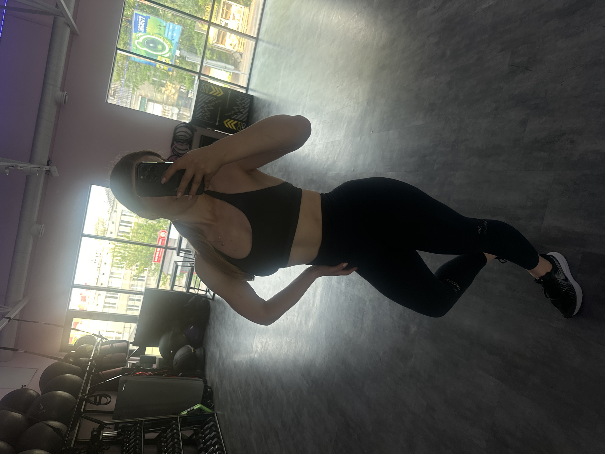 The image captures a full-body mirror selfie taken by a woman in a gym setting in Ternopil, Ukraine. The photograph is oriented 90 degrees clockwise from a standard upright view.
The main subject is a woman, seen from approximately the chest down to her feet. She is wearing black athletic attire, consisting of a sports bra and form-fitting leggings with subtle patterned cutouts near the ankles. She has black athletic shoes with white soles. Her right arm is bent, holding a smartphone in front of her face, which obscures her facial features. Her left hand rests on her hip, and a delicate necklace is visible around her neck. Her pose suggests a momentary pause during or after a workout.
The setting is an indoor gym characterized by a dark, possibly concrete or tiled, floor and light-colored walls, possibly light pink or purple in areas. Large, multi-pane windows dominate the background, allowing bright natural light to flood the space, indicating it is daytime. Outside the windows, a vibrant city scene of Ternopil is visible, featuring multi-story buildings, lush green trees, and glimpses of a street.
Within the gym, various pieces of equipment are present. To the left (when correctly oriented), a rack holds several large, dark medicine balls. Further left, a rack of dumbbells is partially visible. Other gym apparatus, including what appears to be a workout bench, bands, and bags, are also scattered around.
Visible text includes a prominent sign outside the windows, featuring the word "SPORT" in white letters on a green background, along with a circular logo. Other yellow and black geometric patterns or signs are seen on the wall or equipment covers near the windows. The bright conditions outside suggest a sunny day. The overall impression is a modern, well-lit fitness facility.