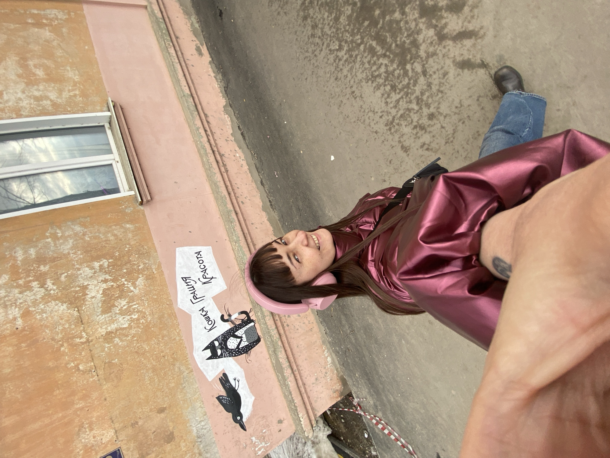 A medium shot captures a person standing beside a building in Votkinsk, Russia. The person is wearing pink headphones and a shiny, maroon-colored jacket, and jeans. They appear to be taking a selfie, with a portion of their arm and hand visible in the foreground. They are smiling towards the camera. Beside them, on the wall of a building, is a mural featuring a black cat and a black bird. The text "Кошка Грация красота" is written in Cyrillic beside the cat, meaning "Grace cat beauty". Above the mural, there's a window with a white frame, and the building is painted in various shades of pink and orange. The ground is visible, appearing to be a paved area or sidewalk.