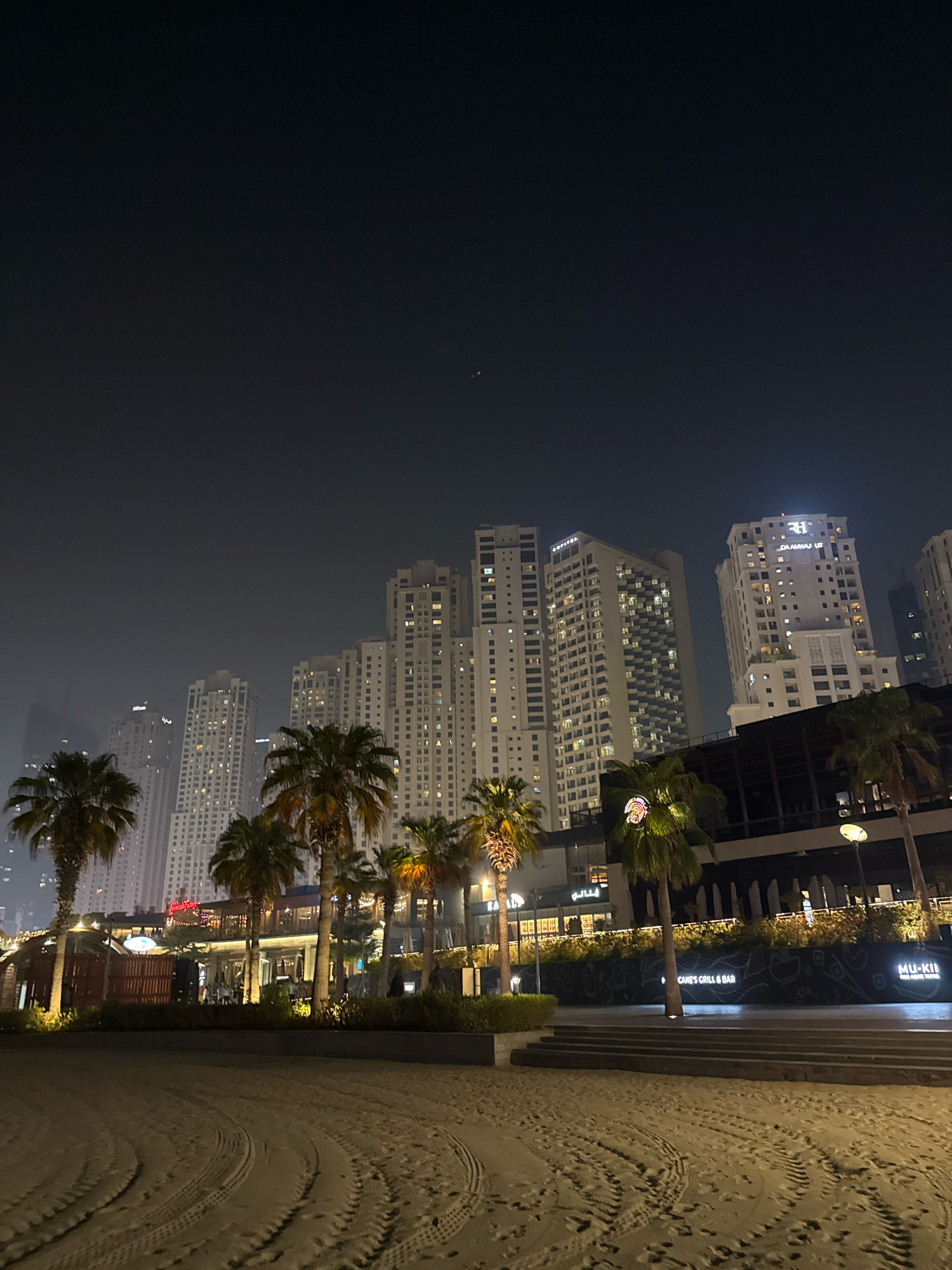 This is a nighttime photograph of the Jumeirah Beach Residence (JBR) waterfront in Dubai, United Arab Emirates. The scene features a sandy beach in the foreground with tire tracks from vehicles. Behind the beach, a strip of greenery and a promenade are visible, with several palm trees lining the area. Further back, a row of illuminated buildings, including high-rise apartments and hotels such as the Sofitel and the Hilton, are prominent against the dark sky. Restaurants and shops are visible on the ground level of some of these buildings, with neon signs and outdoor seating areas. The overall atmosphere is one of a bustling urban waterfront at night.