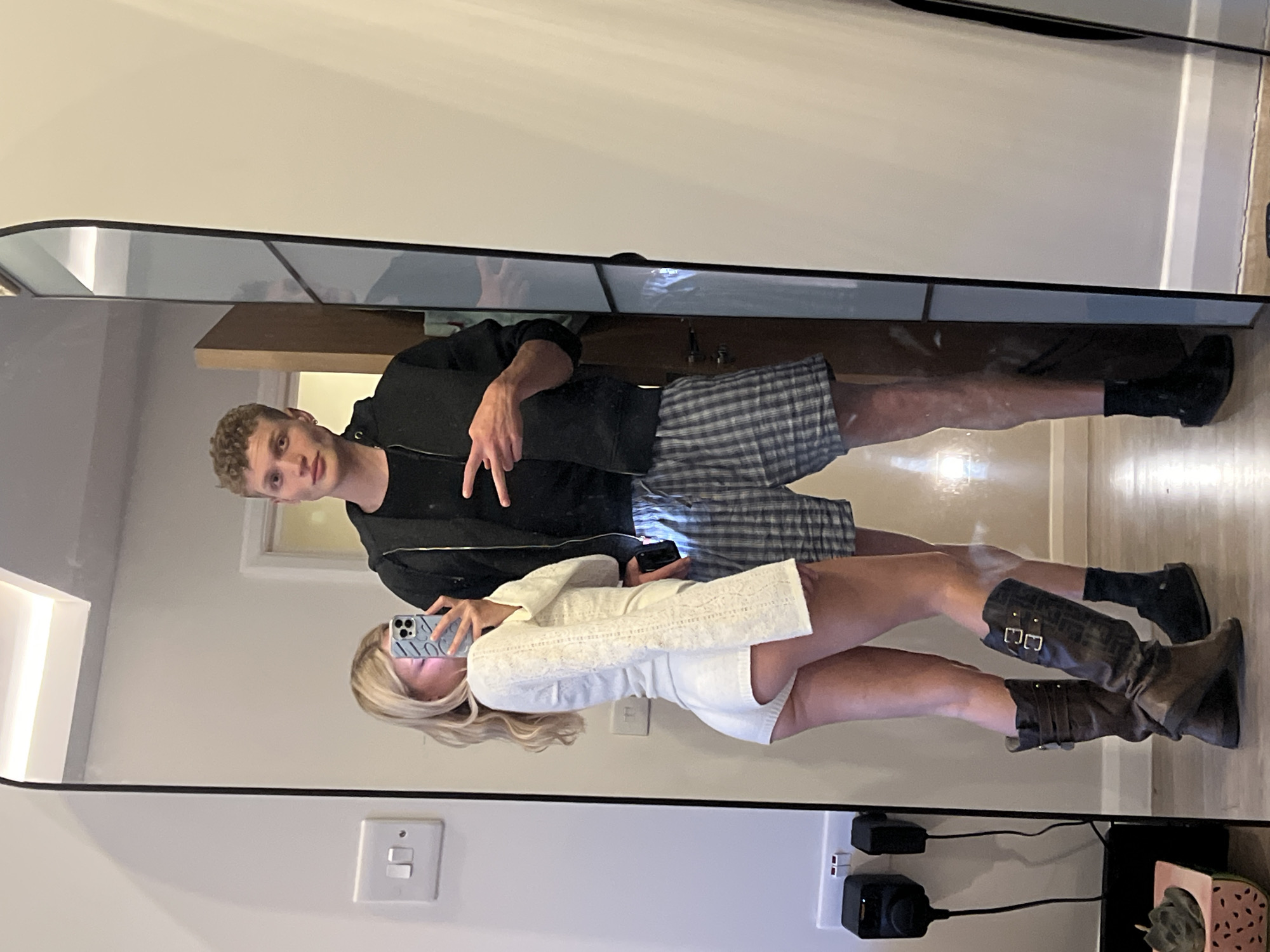 A man and a woman are taking a mirror selfie in a hallway. The woman is on the left, wearing a white textured romper and brown knee-high boots. She is holding a phone up to take the picture, her face partially obscured by it. The man is on the right, wearing a black zip-up hoodie and plaid boxer shorts. He is making a peace sign with his right hand. The background shows a light-colored wall, a door, and a large mirror. There are electrical outlets and a black speaker on the wall to the left. The floor is wooden. The scene appears to be indoors, likely in a residential setting, but there is no clear indication of its specific location within London, United Kingdom.