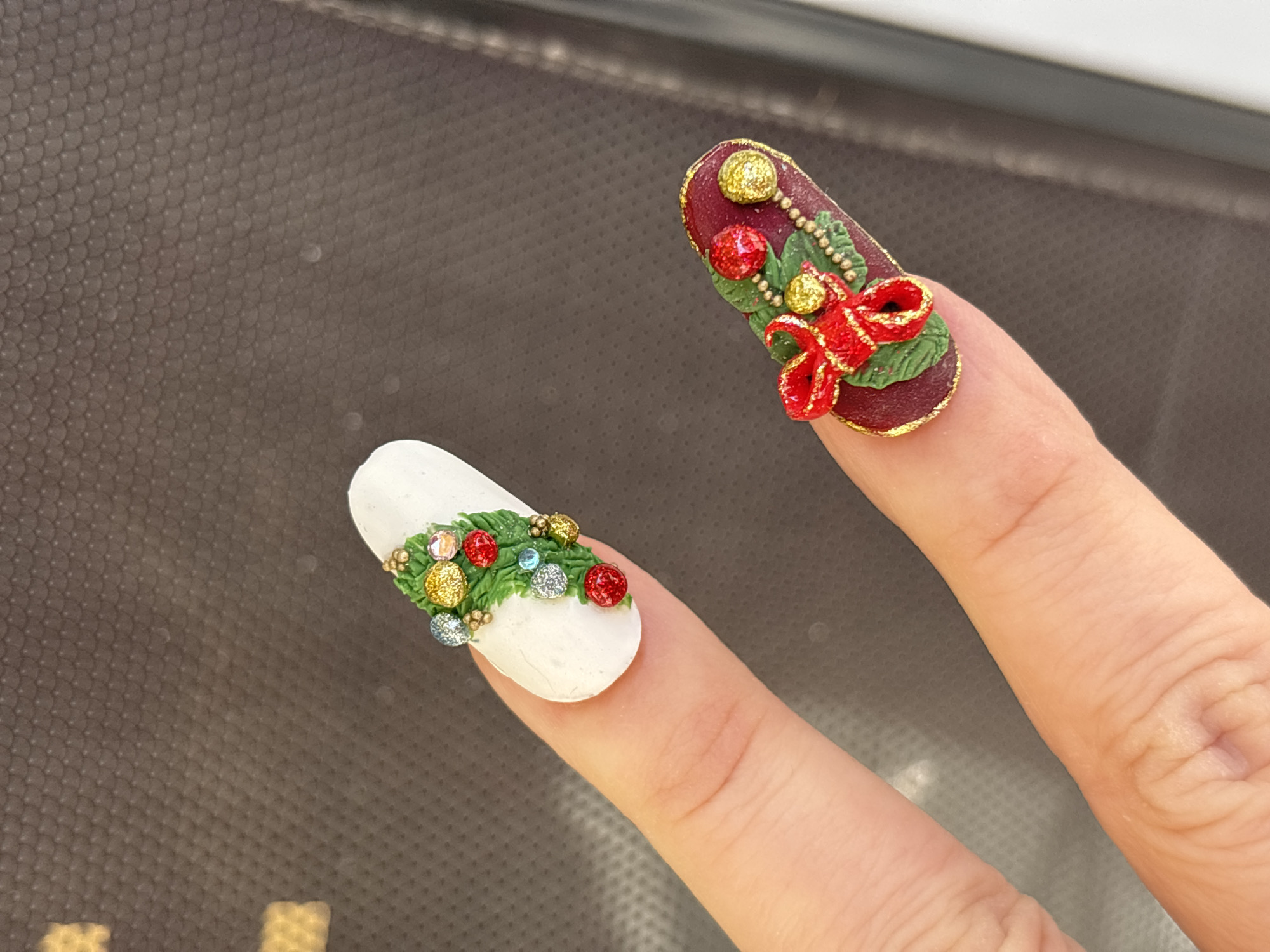 The image presents a close-up view of two fingers displaying two elaborately decorated artificial nails, highlighting intricate, festive-themed nail art. The fingers are positioned such that one nail appears above and slightly behind the other, held up for clear visibility.
The top nail is long and oval, featuring a rich, dark red (burgundy) base with a shimmering gold glitter border. Adorning its surface is a raised, three-dimensional design resembling a Christmas garland. This design incorporates textured green leaves, accented with sparkling red and gold spherical ornaments or "berries," and a prominent red bow with golden glitter details.
The bottom nail, held by a different finger, is also oval-shaped but has a matte white base. It showcases a similar raised decorative element of green foliage, intermingled with various glittery spheres in shades of red, gold, and silver, alongside a few small, clear or light blue rhinestones. Both nails exhibit a highly detailed and textural aesthetic, indicative of specialized holiday nail art.
The background is a dark, textured surface, possibly a patterned fabric or material, exhibiting a fine, repeating scale-like or mesh pattern. A slightly blurred, lighter band runs horizontally across the upper part of the background, suggesting an indoor setting with artificial lighting. The image focuses on the craftsmanship of the nail designs, without revealing any specific location cues, time of day, or weather conditions. No text is visible in the frame.