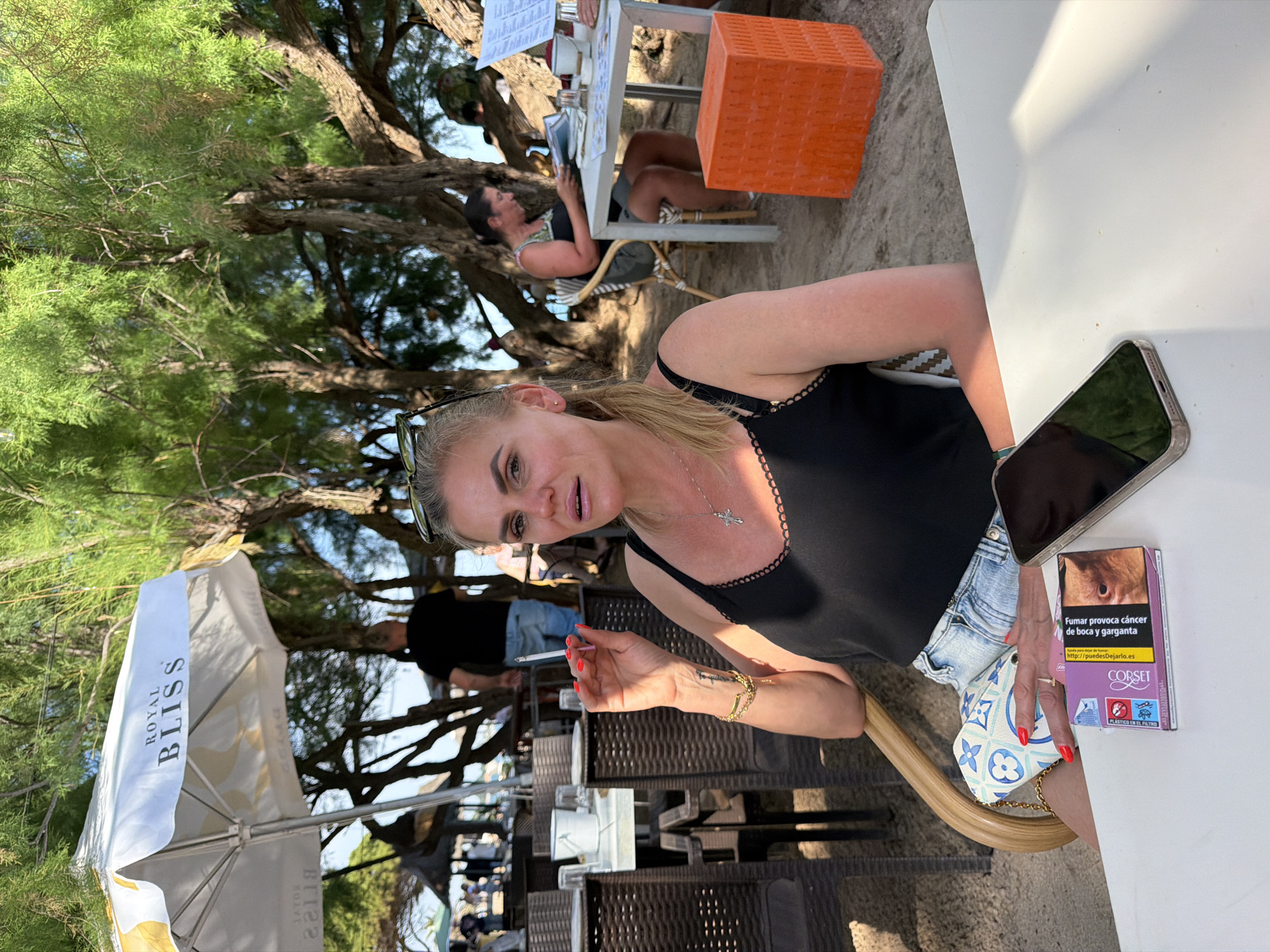 Here is a description of the image:
The image is taken outside at a restaurant or cafe, likely in Mont-roig del Camp, Spain. A woman with blonde hair is seated at a white table and is holding a lit cigarette. She is wearing a black tank top and light-colored denim shorts. On the table is a pack of "Corset" cigarettes in Spanish, a cell phone, and a small Louis Vuitton purse. In the background, other patrons are seated at tables under the shade of trees. There is a white beach umbrella labeled "Royal Bliss" nearby.