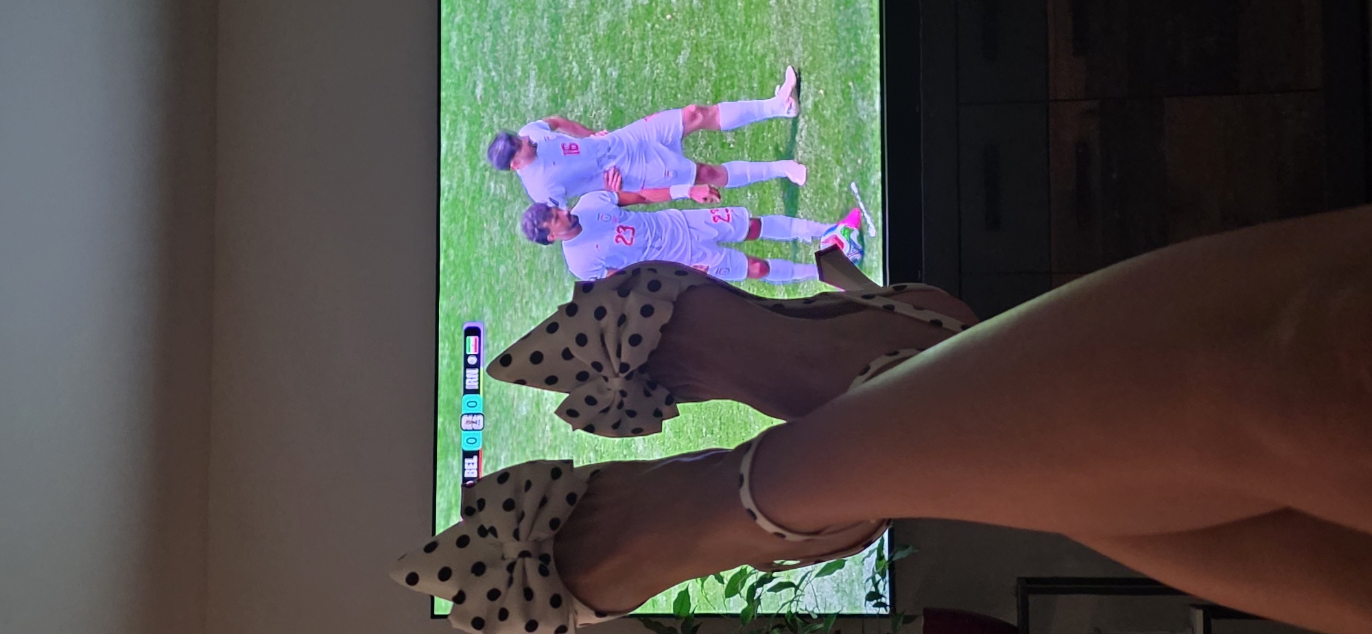 The image shows a person's legs, adorned with polka-dotted high heels, raised in front of a television screen. The television displays a soccer match between Belgium (BEL) and Iran (IRN), with the score currently 0-0. Two male soccer players in white jerseys are visible on the screen, one wearing the number 23 and the other number 16. The setting appears to be a living room, with a dark cabinet visible below the television. The scene is dimly lit, with the primary light source coming from the television. The image does not contain any information about the city of Lieli, Switzerland.