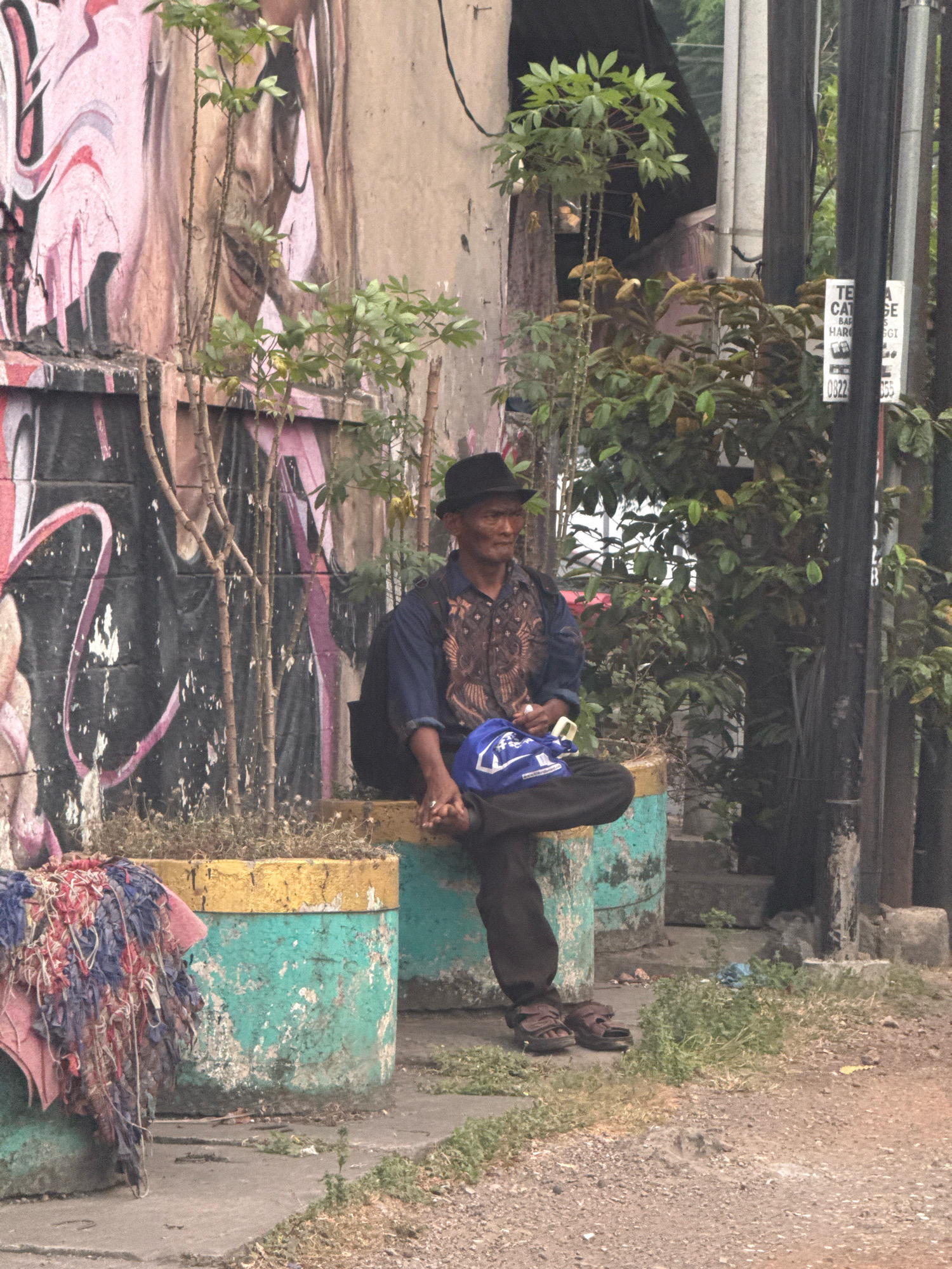 An adult male is seated on a planter box outdoors, possibly in Surabaya, Indonesia, given the context. He wears a black fedora hat, a blue patterned shirt, and dark pants. His legs are crossed, and he holds a blue bag. The planter box he sits on is weathered and painted in teal and yellow. Behind him, a wall is covered in graffiti and several slender plants grow from the planter. To the right of the man, a sign is visible on a pole. The ground in front of the planter is paved with concrete and gravel, with patches of dry grass. A pile of colorful, tattered fabric is draped over another planter to the left.